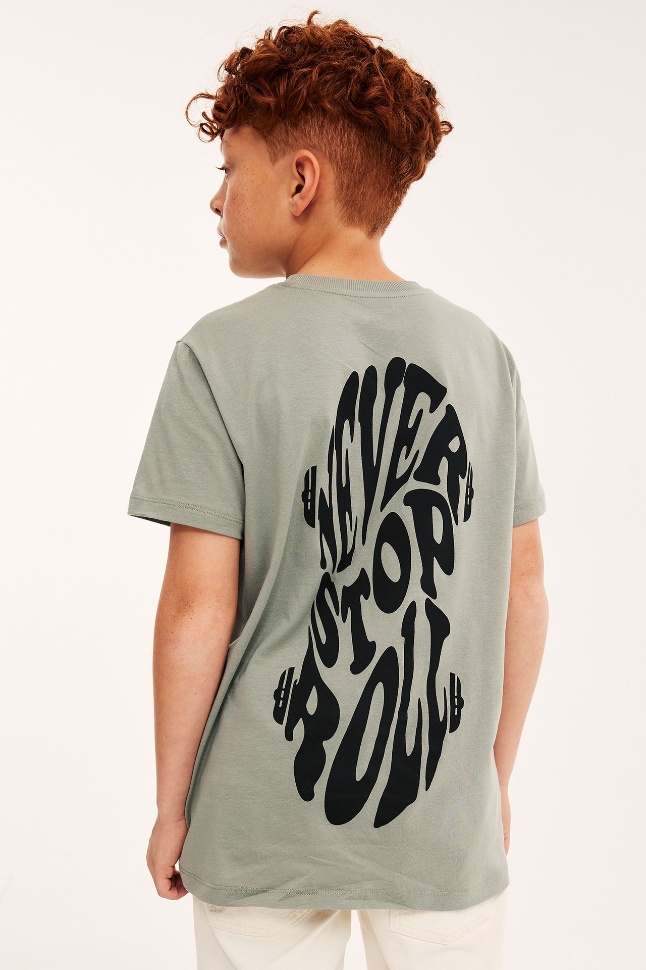 Dziecko, widok z tyłu: ciemnozielony bawełniany T-shirt z dużym czarnym nadrukiem "NEVER STOP ROLLIN'', jasne spodnie.