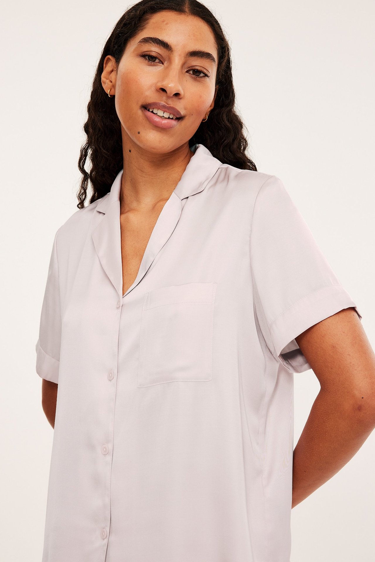 Lähikuva edestä: nainen vaaleanpunaisessa satiinisessa lyhythihaisessa pyjamapaidassa, jossa kaulus ja rintatasku.