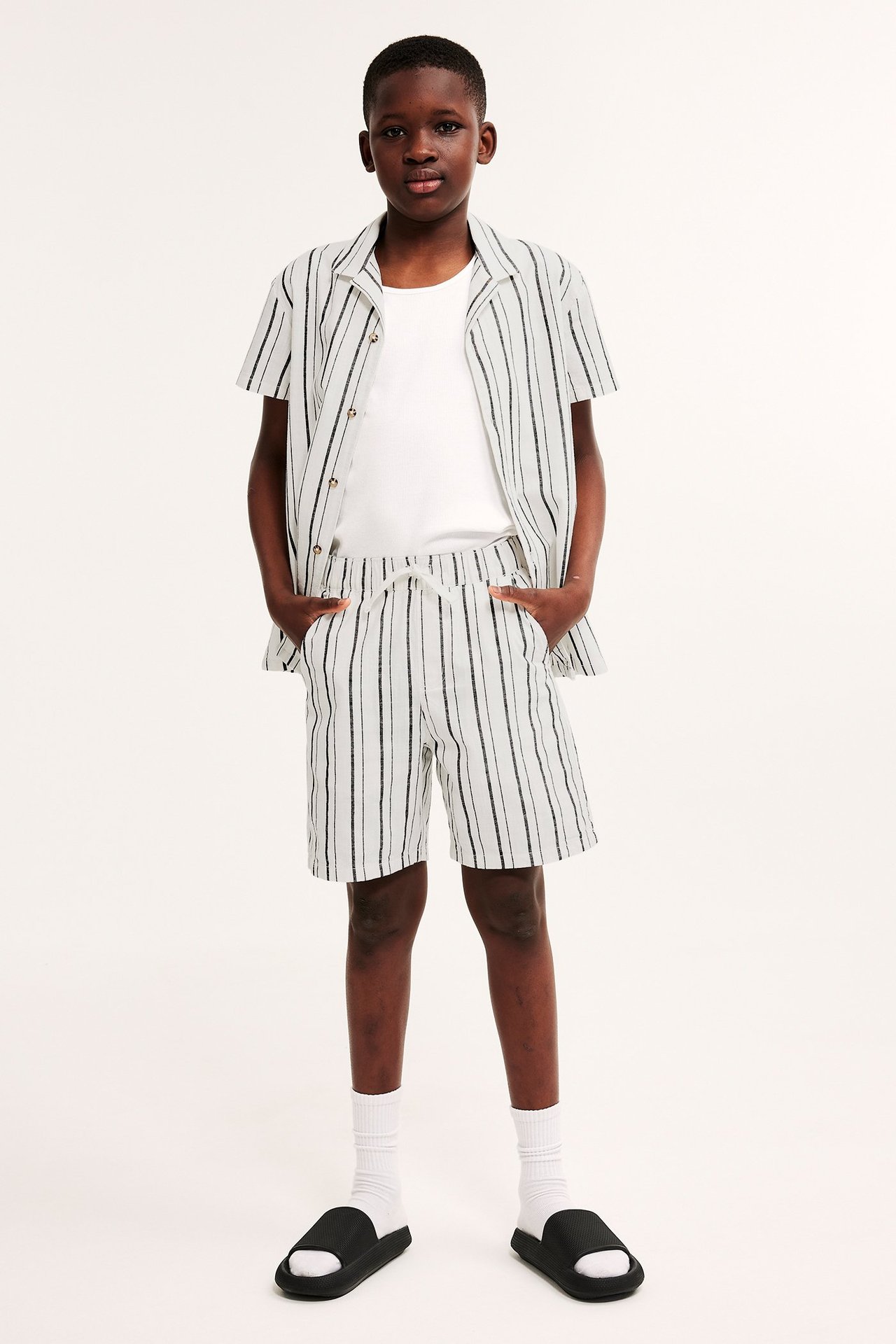 Widok z przodu: Biała koszula w paski, szorty, biały T-shirt, skarpetki, czarne klapki na dziecku.