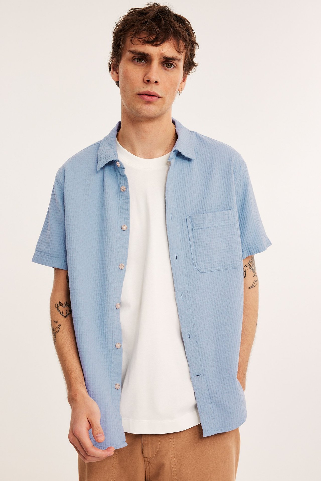 Edestä: mies avoimessa vaaleansinisessä strukturoidussa lyhythihaisessa puuvillapaidassa, valkoisen t-paidan ja ruskeiden housujen päällä.
