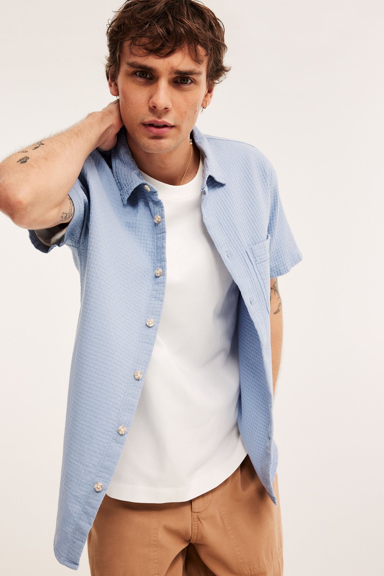 Edestä kuvattu mies yllään avoin vaaleansininen lyhythihainen paitapusero valkoisen t-paidan ja ruskeiden shortsien päällä.