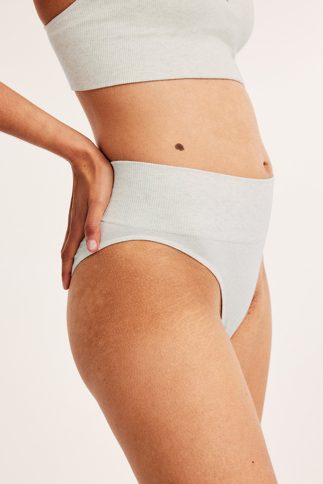 Naisen sivukuva vaaleanharmaissa meleeratuissa saumattomissa ribatuissa Brazilian-alushousuissa ja yhteensopivissa rintaliiveissä.