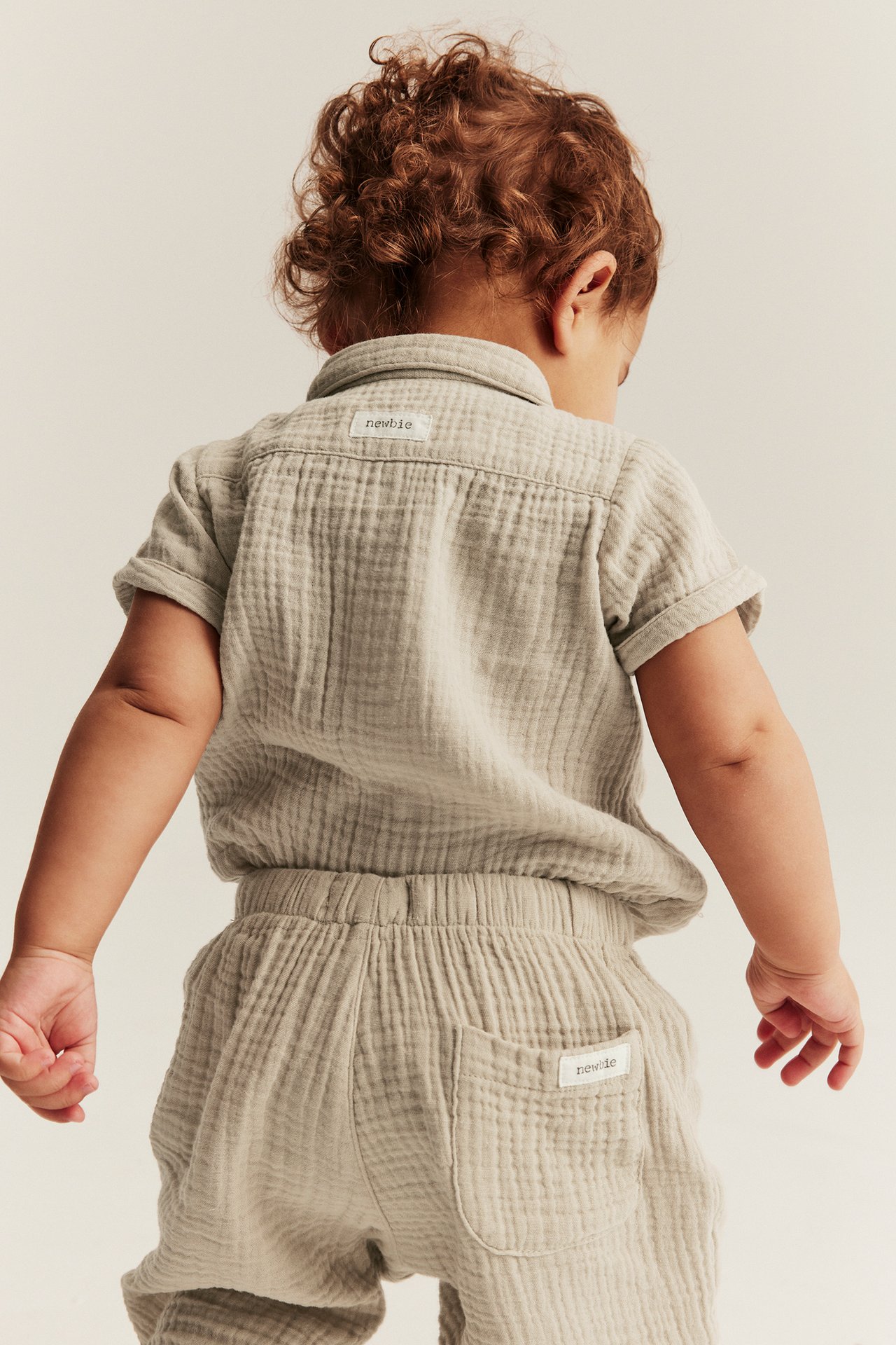 Bakfra av baby i beige kortermet bomullsskjorte og matchende bukser med elastisk midje.