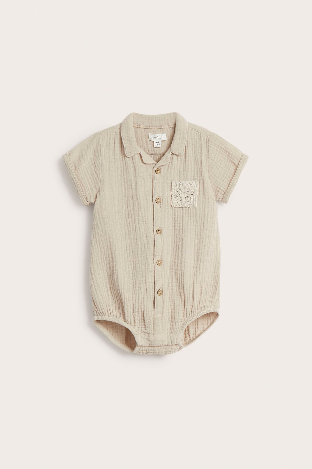Framifrån, beige kortärmad babybody i bomullsmuslin med knappar och en texturerad bröstficka.