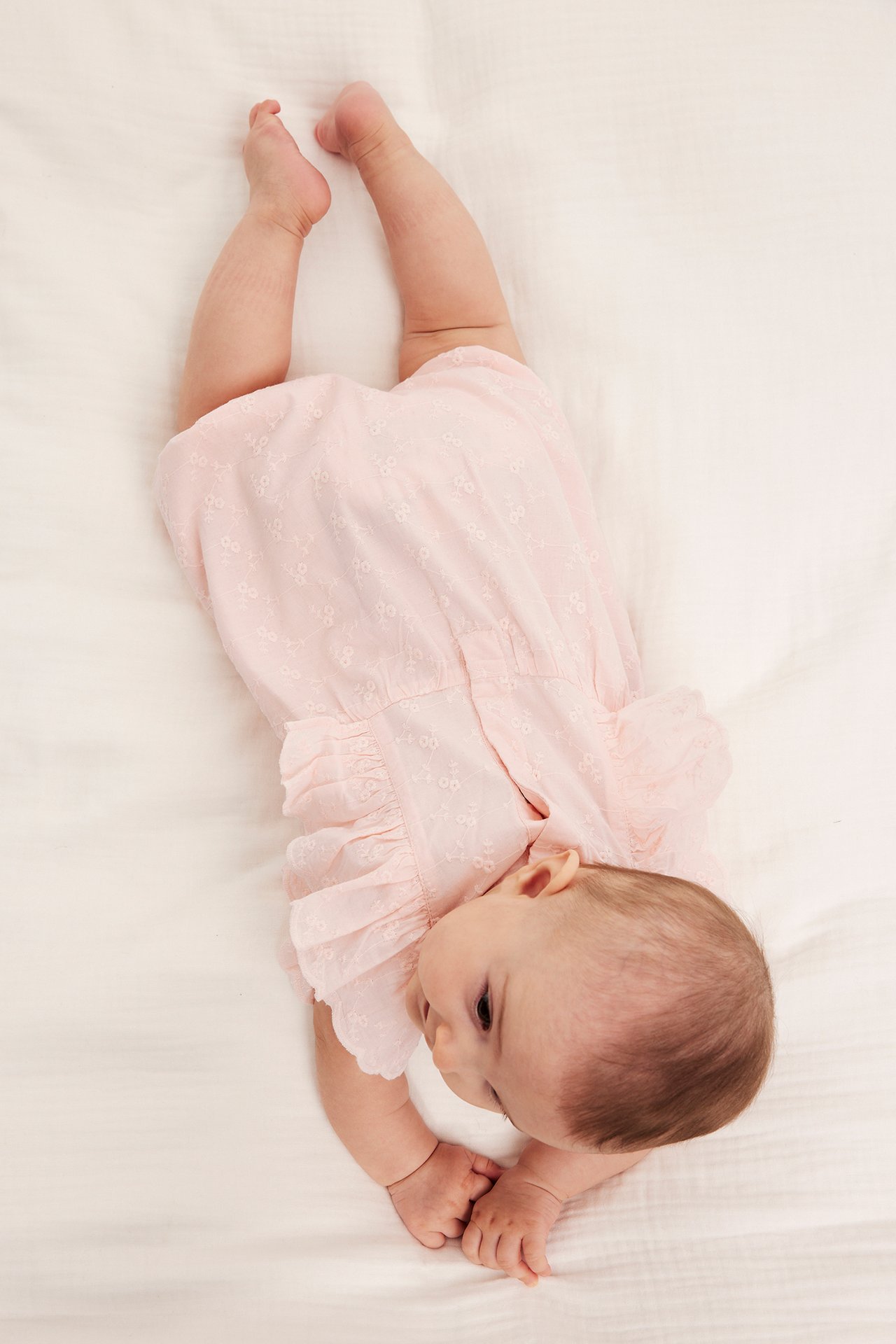 Ovenfra: baby i lyserosa broderie anglaise playsuit med skulderfriller og trykknapper.