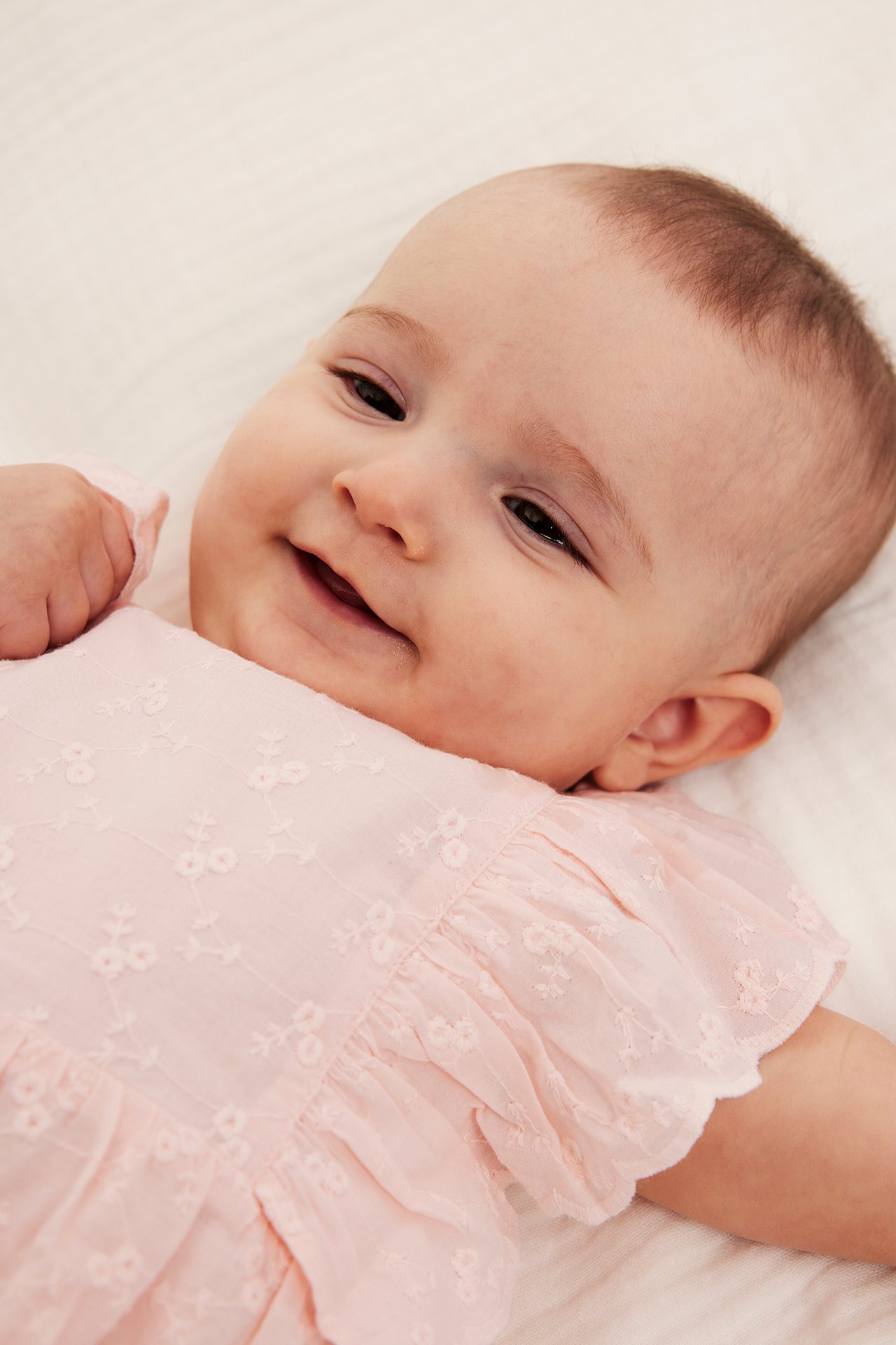 Nærbilde av en smilende baby i lys rosa broderie anglaise playsuit med volangermer.