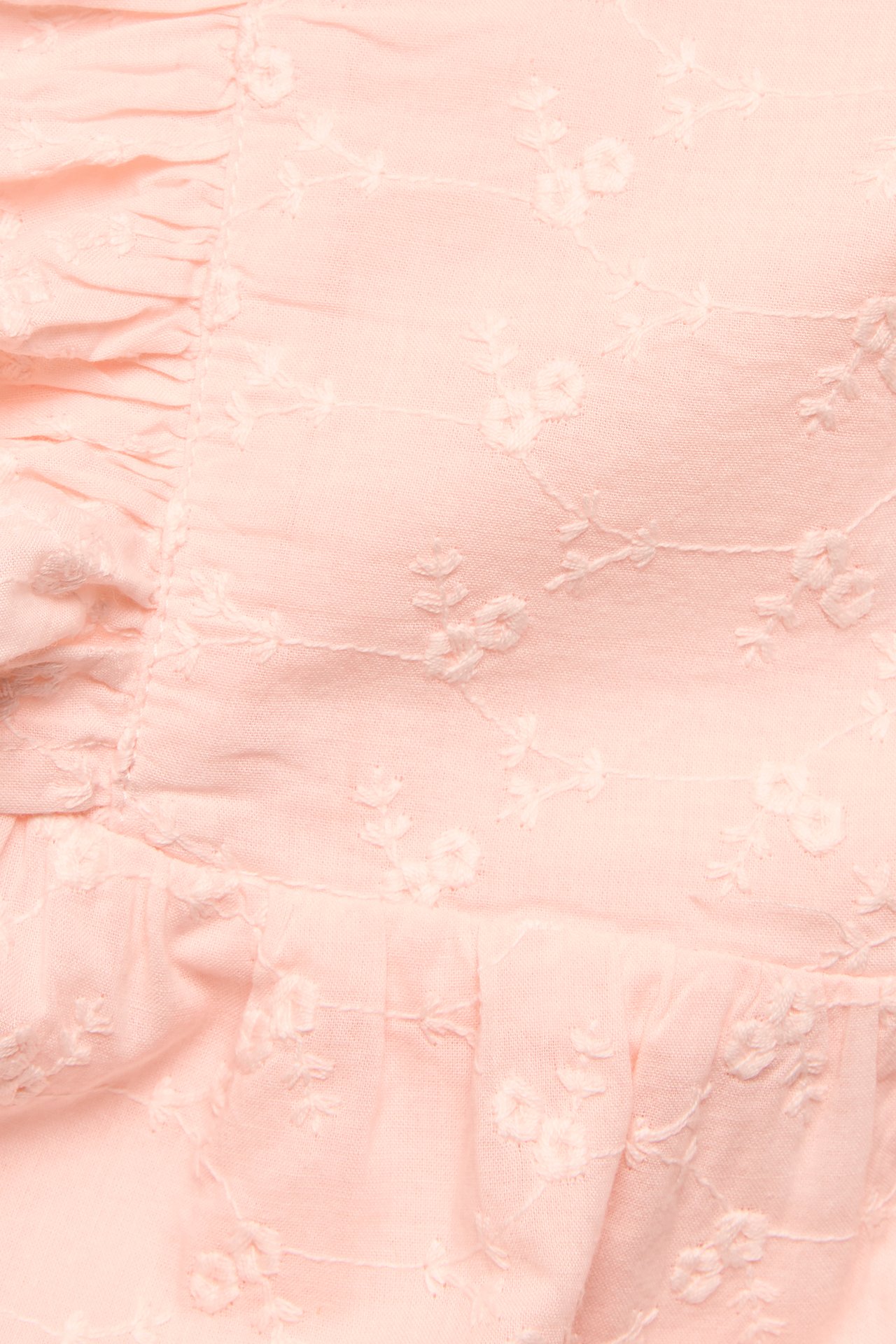 Nærbilde av en lyserosa babyplaysuit med hvit broderie anglaise og volanger.