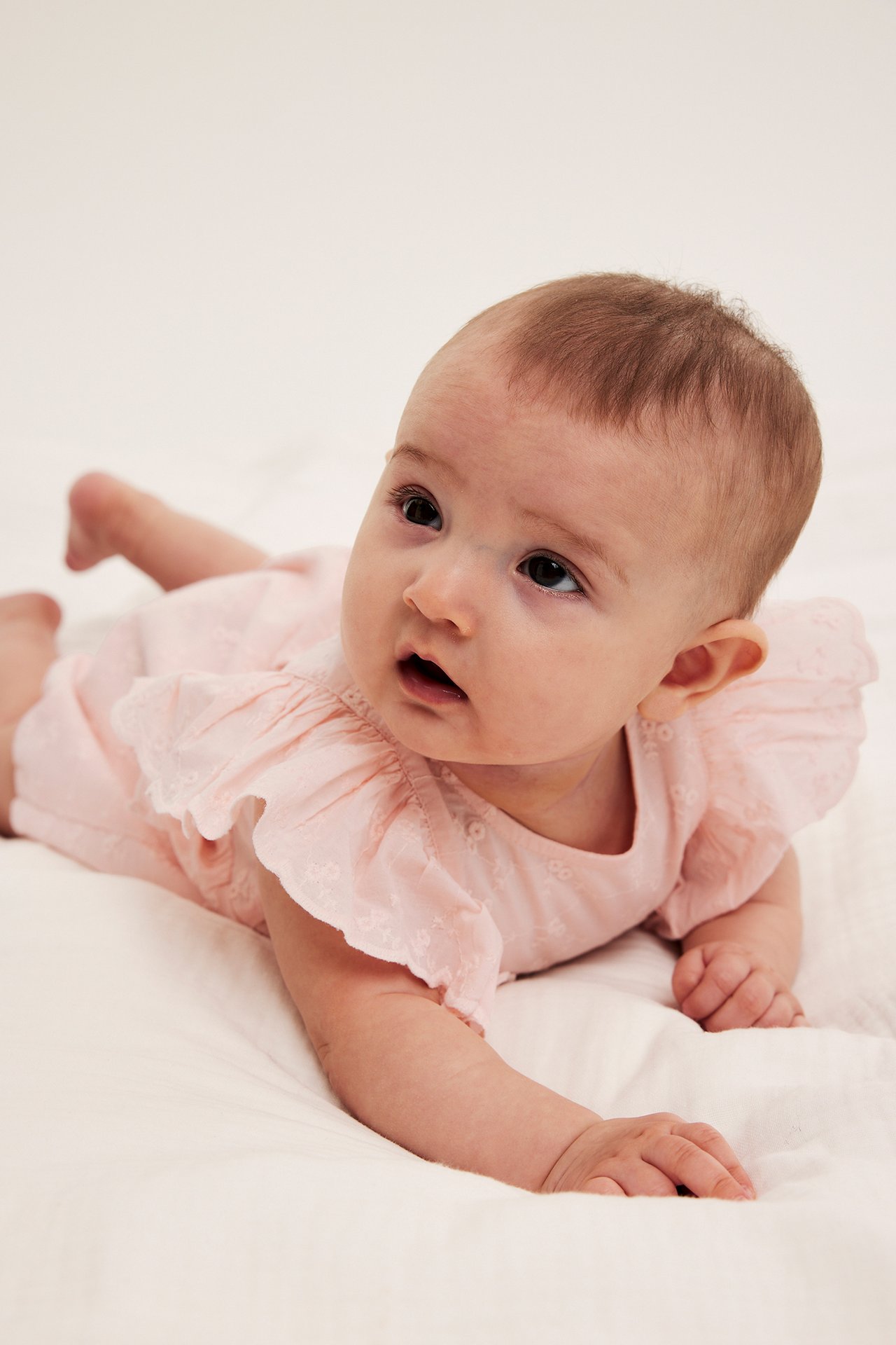 Nærbilde forfra av en baby i lys rosa broderie anglaise playsuit med volangermer.