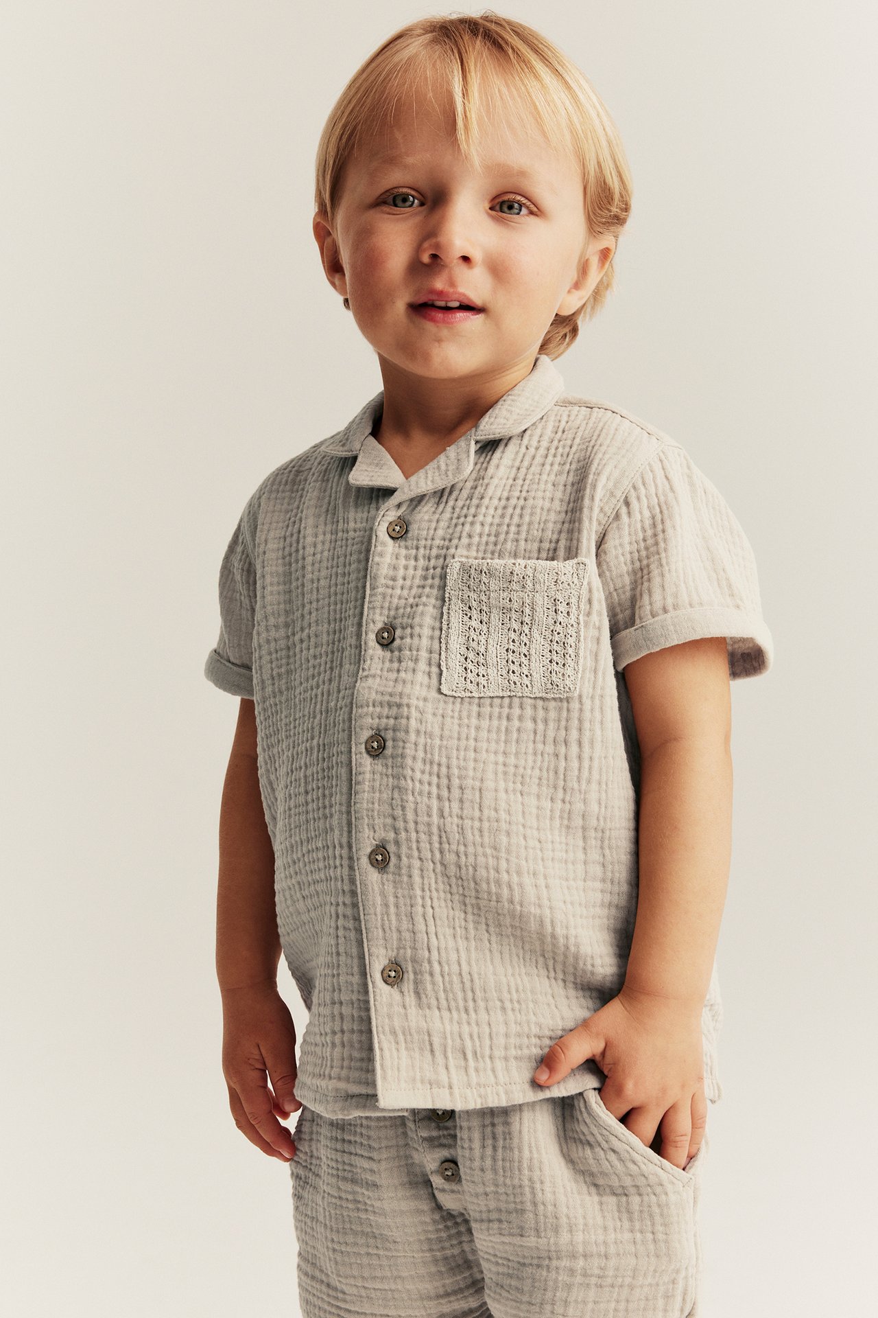 Beige kortärmad skjorta i bomullsmuslin med knappar, texturerad ficka och matchande shorts på ett barn.