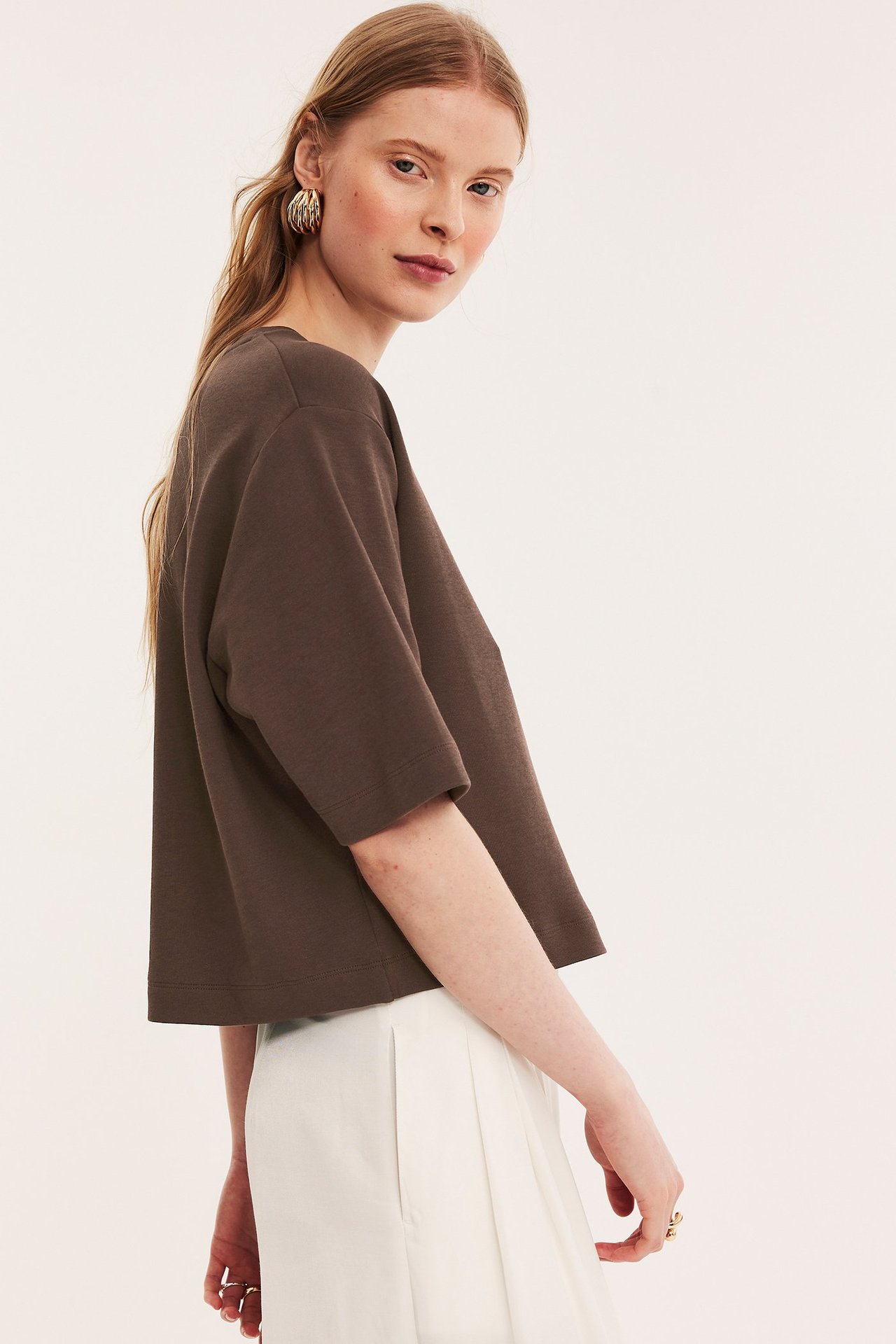 Widok z boku kobiety w ciemnobrązowym, krótkim bawełnianym T-shircie, białych plisowanych spodniach, złotych kolczykach i pierścionku.