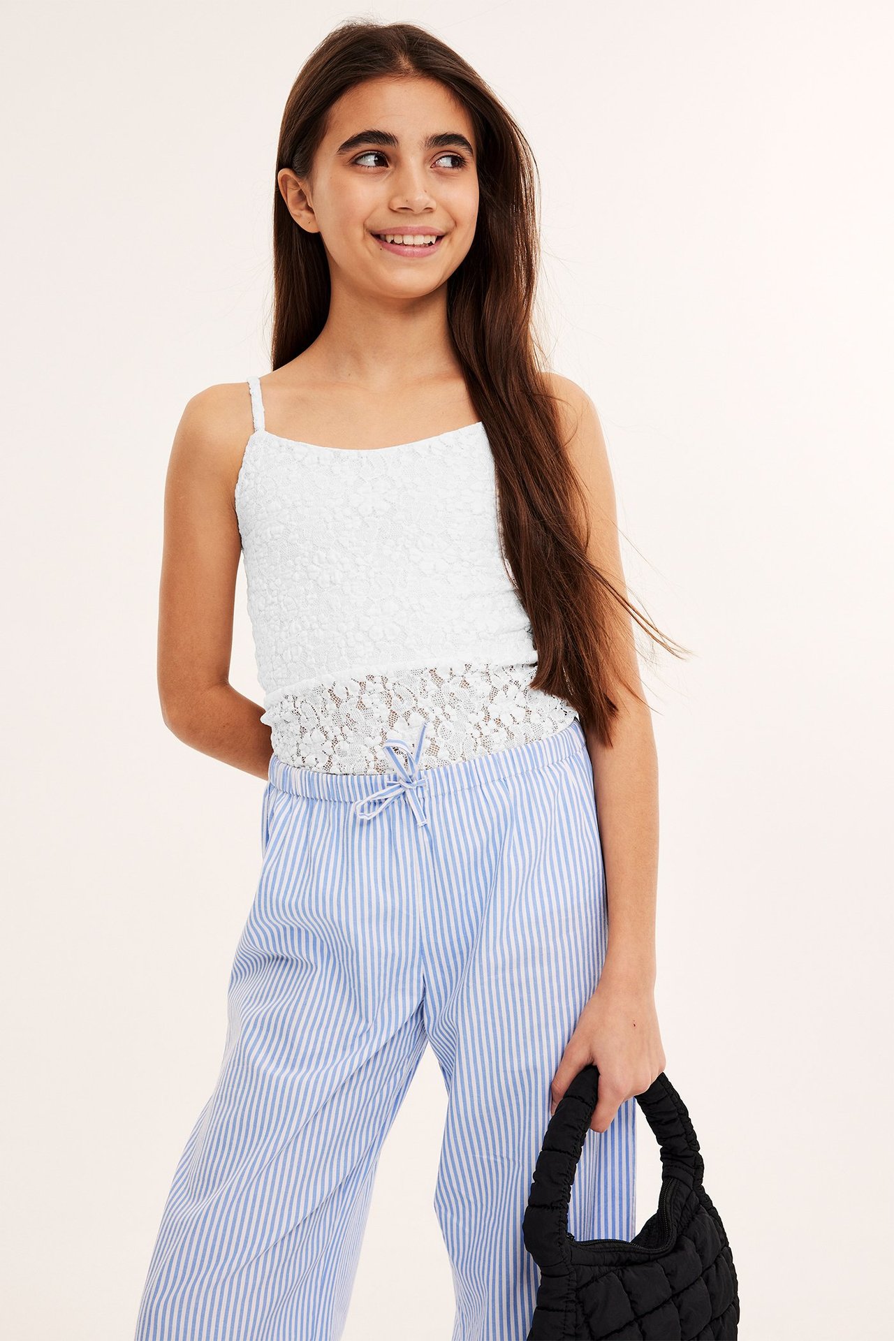 Framifrån, en tjej i vitt spetslinne, blårandiga byxor och håller en svart quiltad handväska.