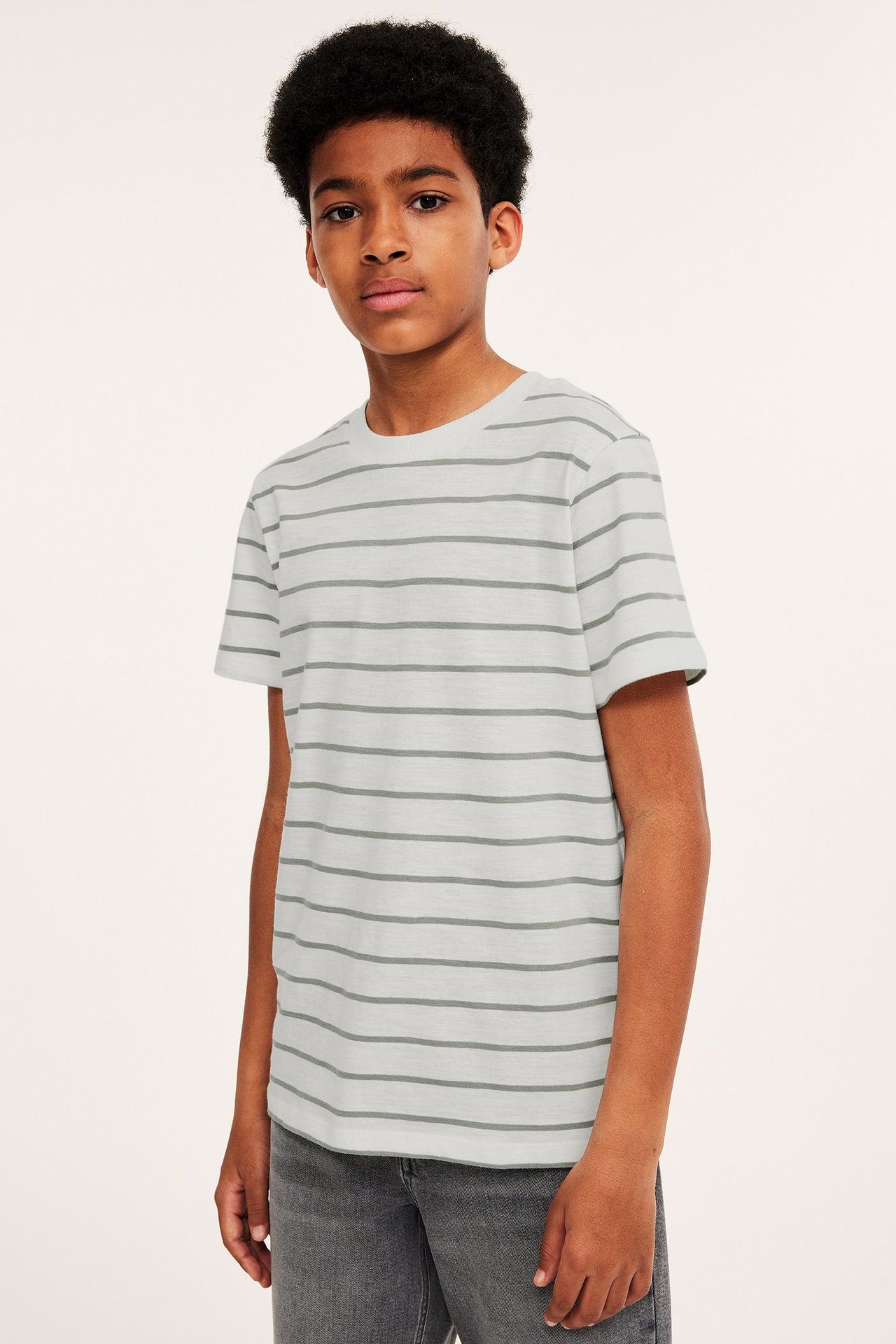 Widok z przodu dziecka w jasnozielonej bawełnianej koszulce w paski z krótkimi rękawami i szarych dżinsach.