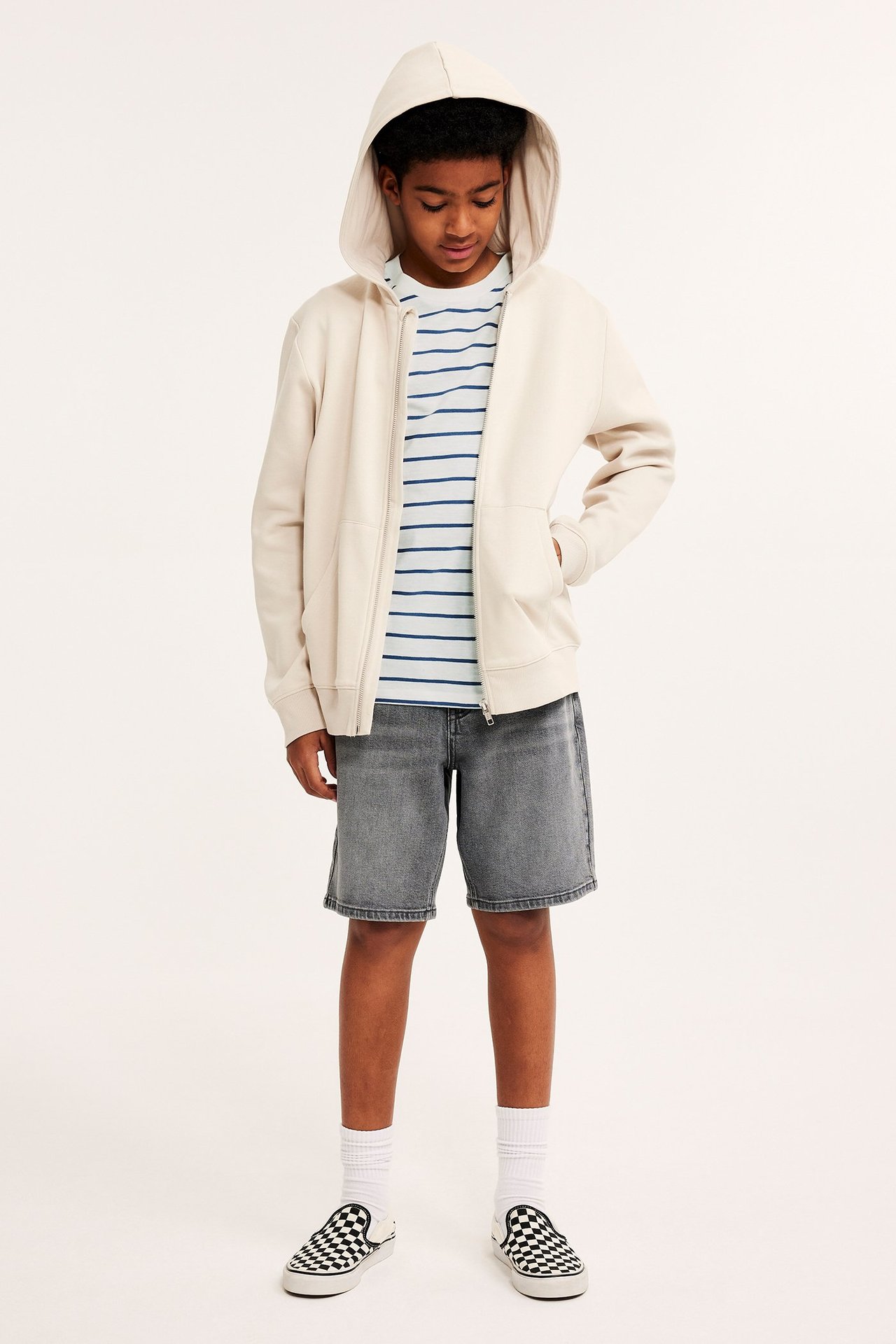 Widok z przodu: Dziecko w niebiesko-białej koszulce w paski, beżowej bluzie, szarych szortach, białych skarpetkach, butach w kratę.