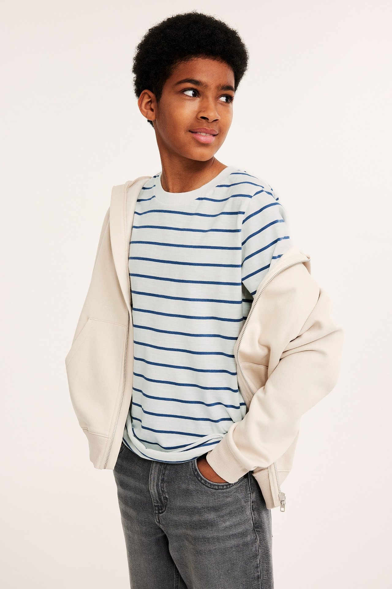 Widok z przodu: dziecko w jasnoniebieskiej bawełnianej koszulce w paski, beżowej rozpiętej bluzie i ciemnoszarych dżinsach.
