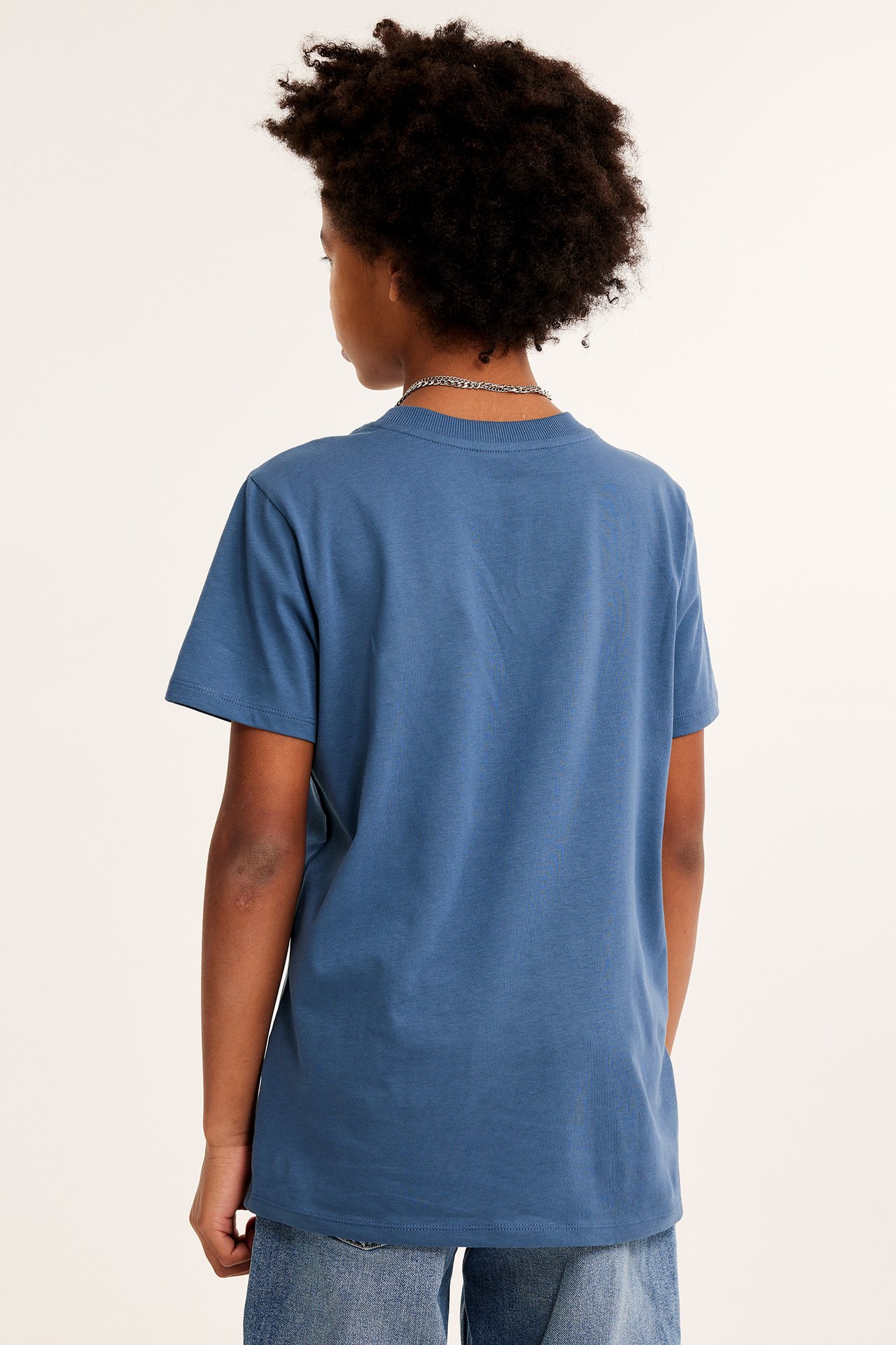 Bakifrån: ett barn i en blå bomulls-t-shirt, blå jeans och en silverkedja.