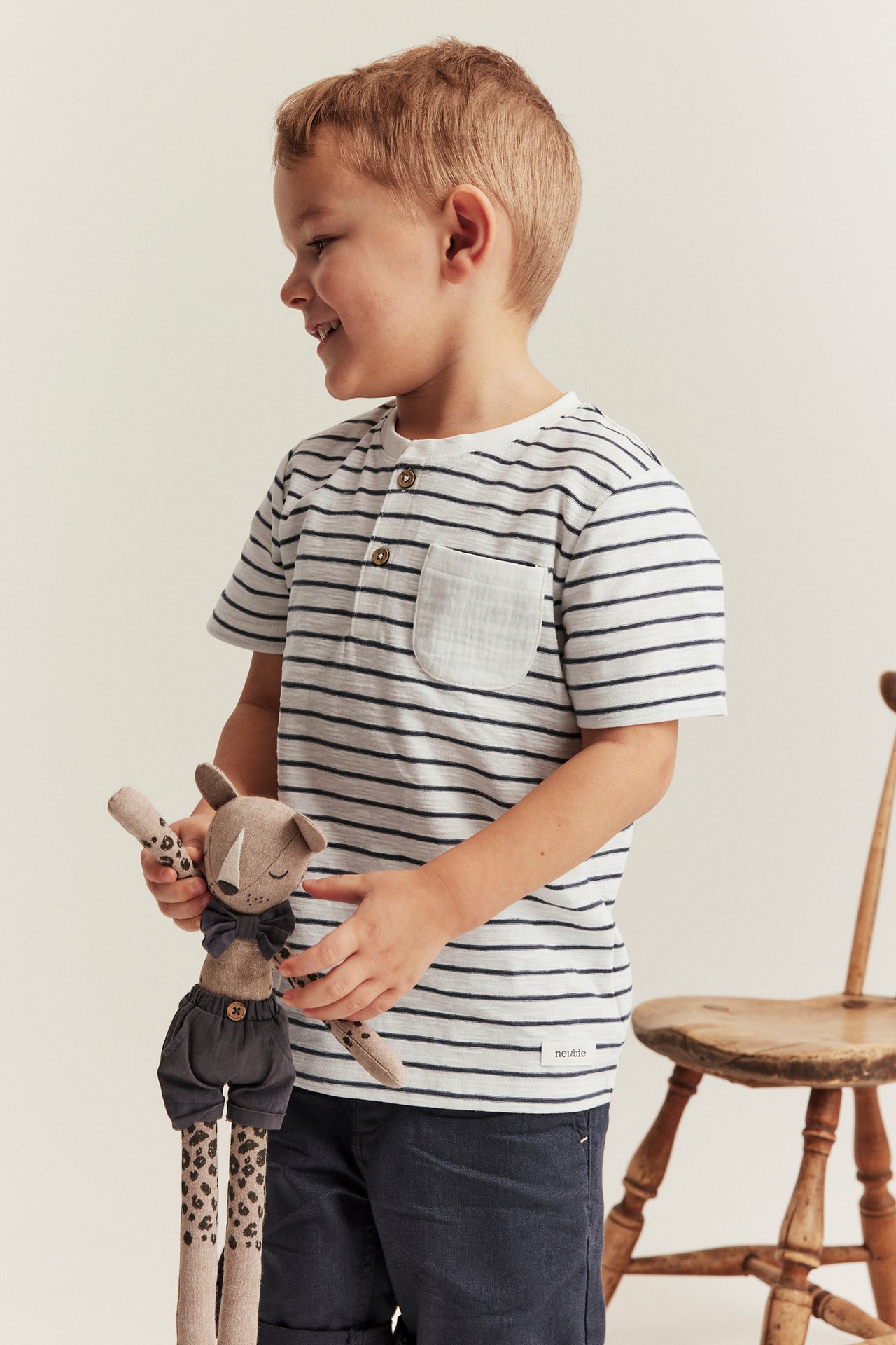 Sidovy: Barn i offwhite randig t-shirt med knappar, mörkblå shorts, håller en brun leksak.