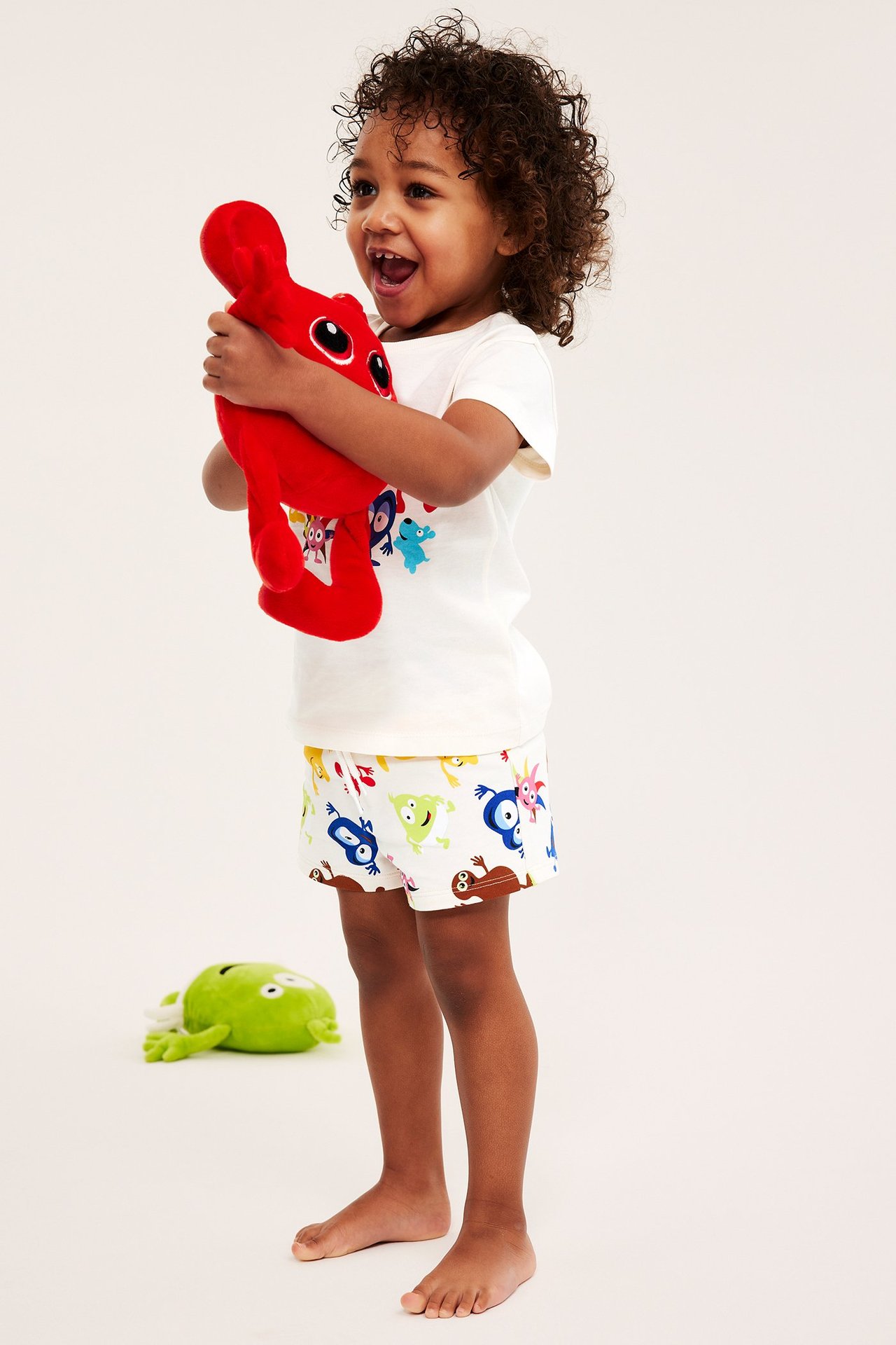 Sivukuva lapsesta, jolla on luonnonvalkoiset Babblarna-kuvioidut shortsit ja t-paita, pitäen punaista pehmolelua.