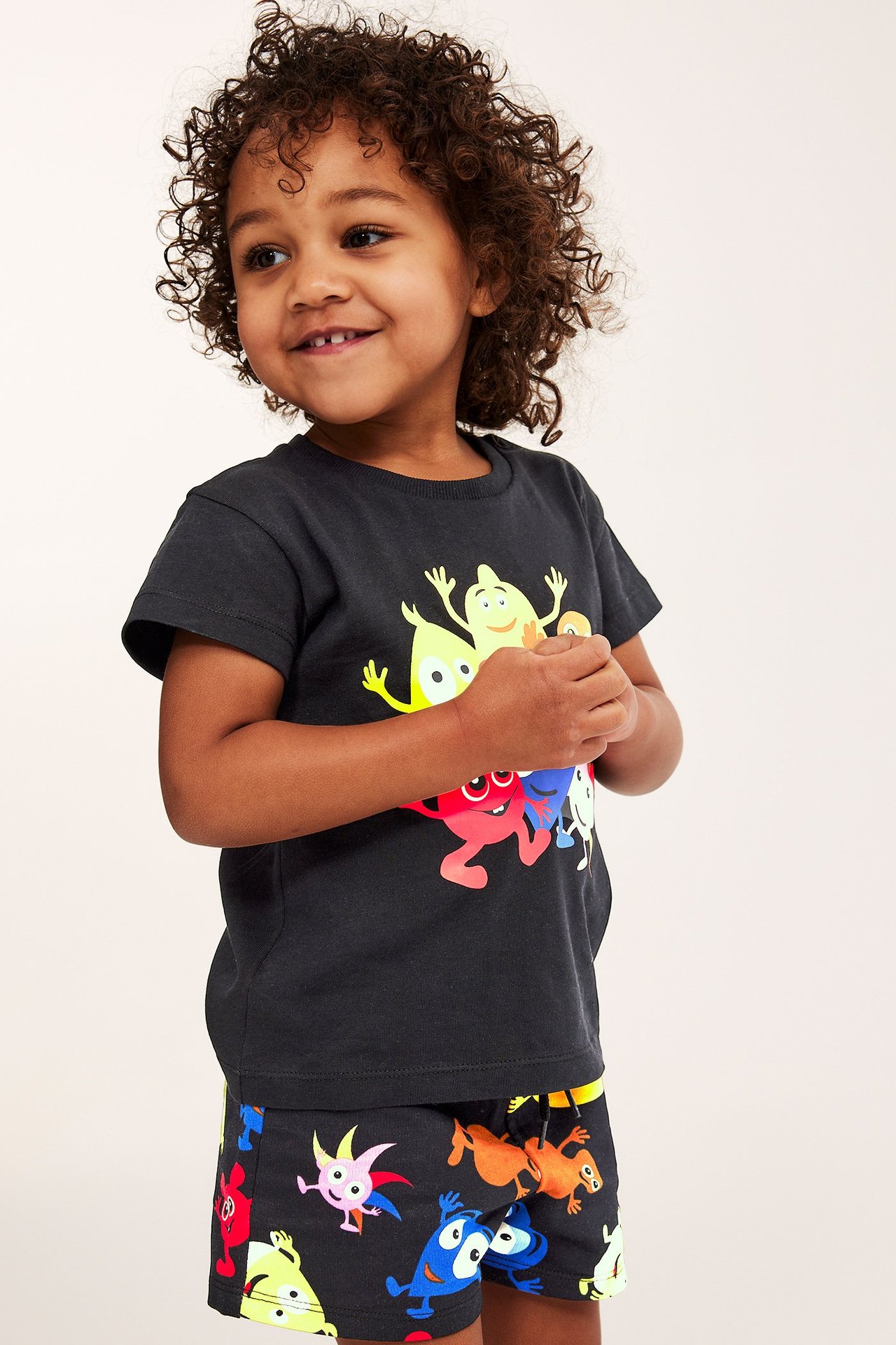 Edestä, hymyilevä lapsi mustissa Babblarna-kuvioiduissa puuvillashortseissa ja yhteensopivassa t-paidassa.