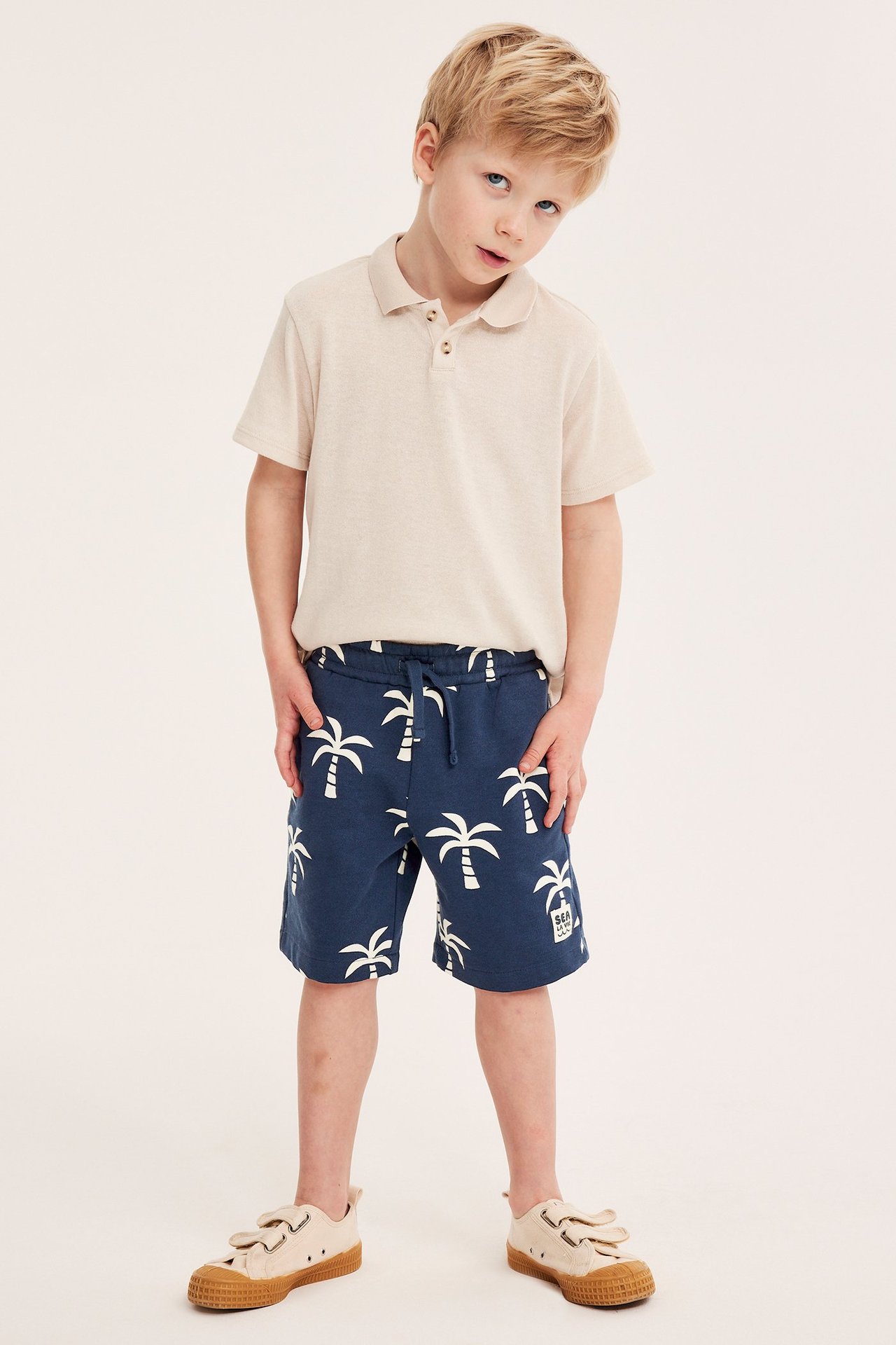 Edestä: lapsi tummansinisissä puuvillaisissa pehmeissä shortseissa valkoisella palmukuvioinnilla, vaaleanbeigessä pikeepaidassa ja beigenvärisissä tennareissa.