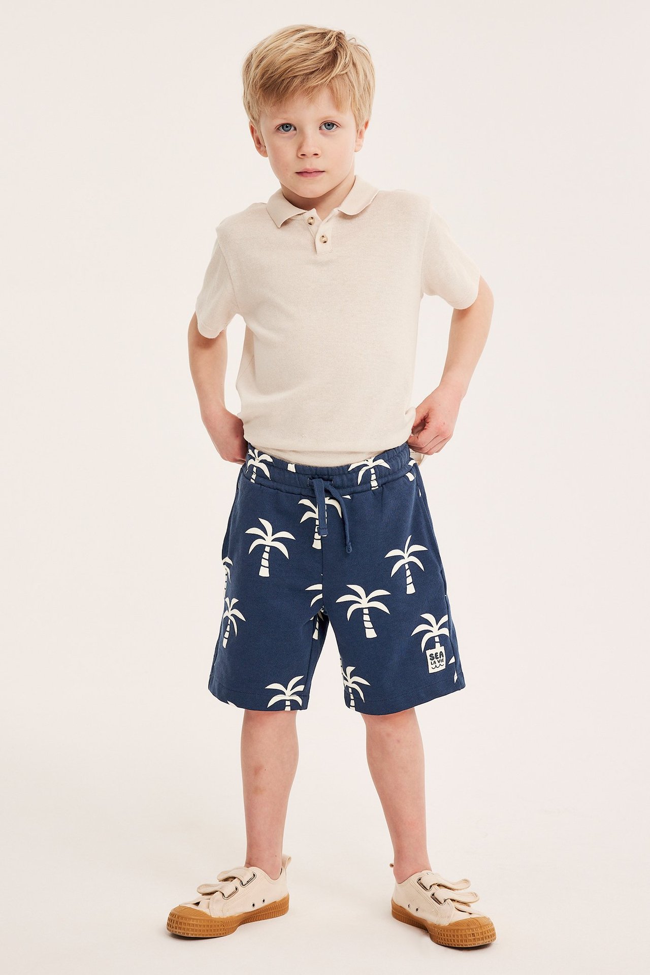 Edestä kuvattu lapsi yllään tummansiniset palmukuvioiset shortsit, beige pikeepaita ja beiget tennarit.