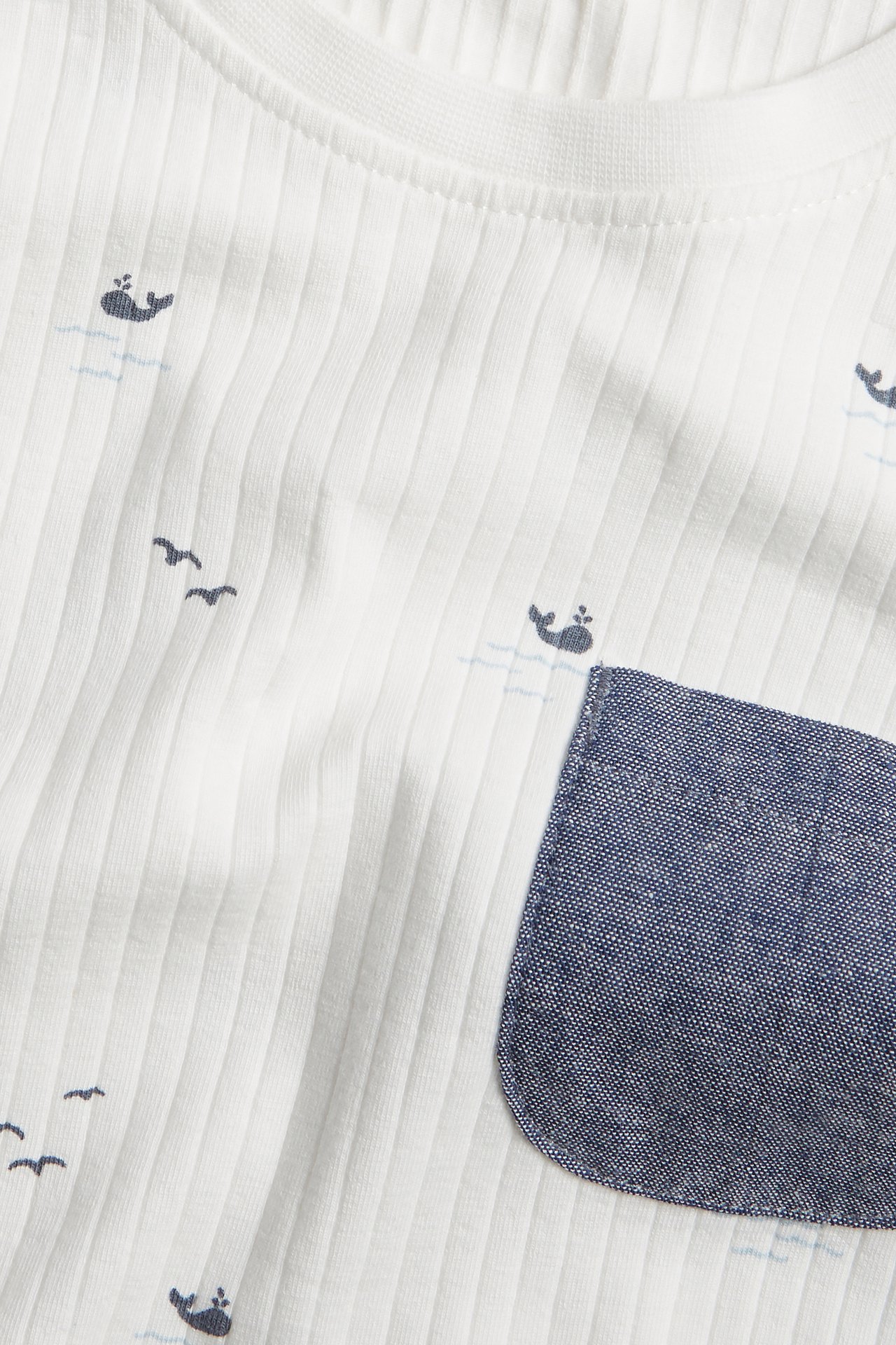 Nærbilde: Offwhite ribbet t-skjorte med blått hvalmønster og mørkeblå chambray-brystlomme.