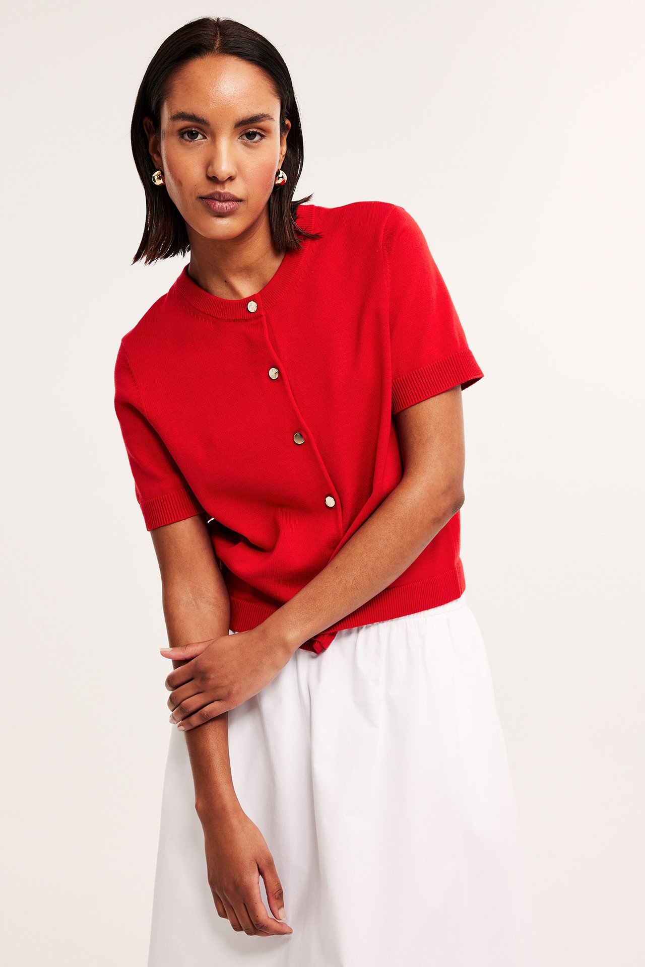 Edestä kuvattu nainen yllään punainen lyhythihainen neuletakki kultaisilla napeilla, valkoinen hame ja kultakorvakorut.