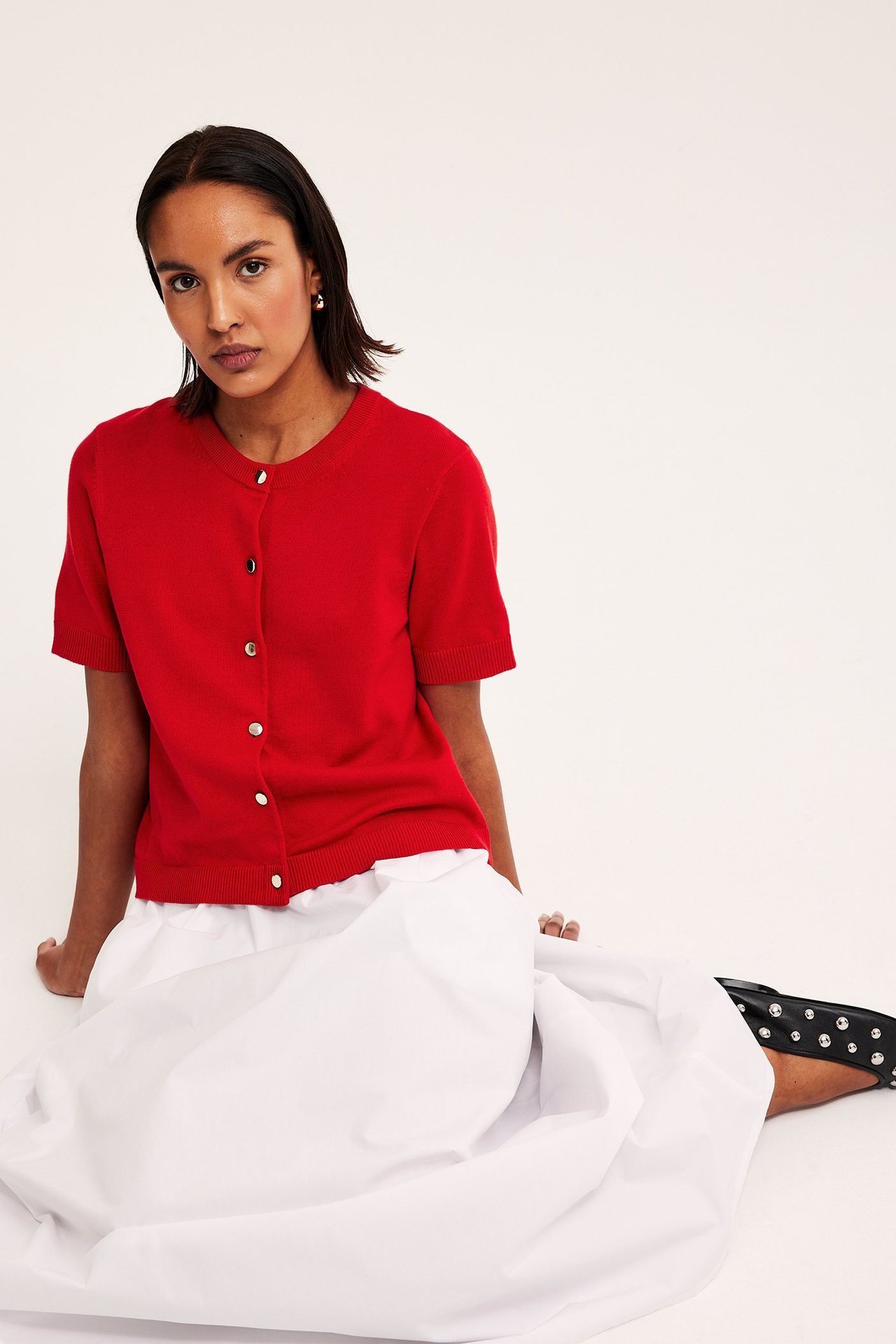 Nainen yllään punainen lyhythihainen neuletakki kultanapeilla, valkoinen hame ja mustat niittikengät.