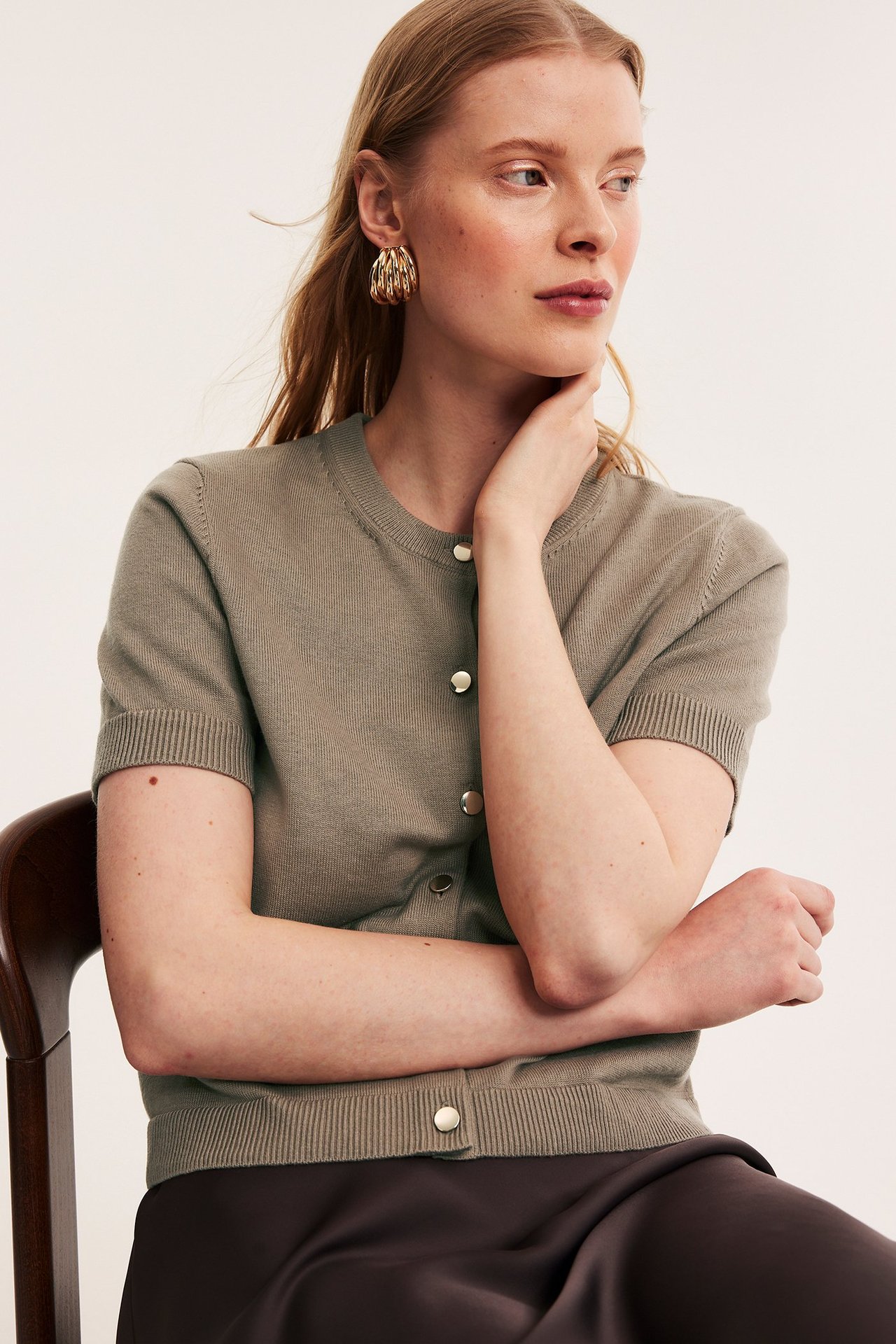 Widok z przodu kobiety w ciemnobeżowym, krótkim kardiganie ze złotymi guzikami, ciemnobrązowej spódnicy i złotych kolczykach kołach.