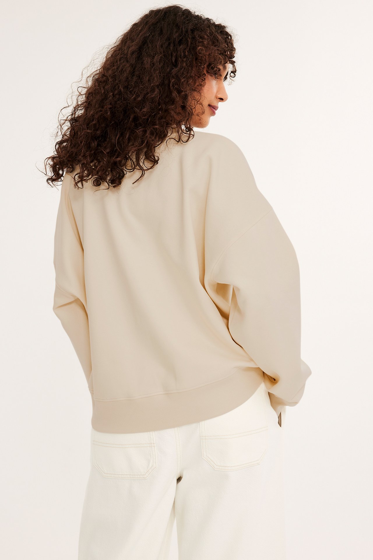 Rygg-/sidevisning av en kvinne i en løstsittende offwhite sweatshirt med ribbet kant og hvite vide bukser.