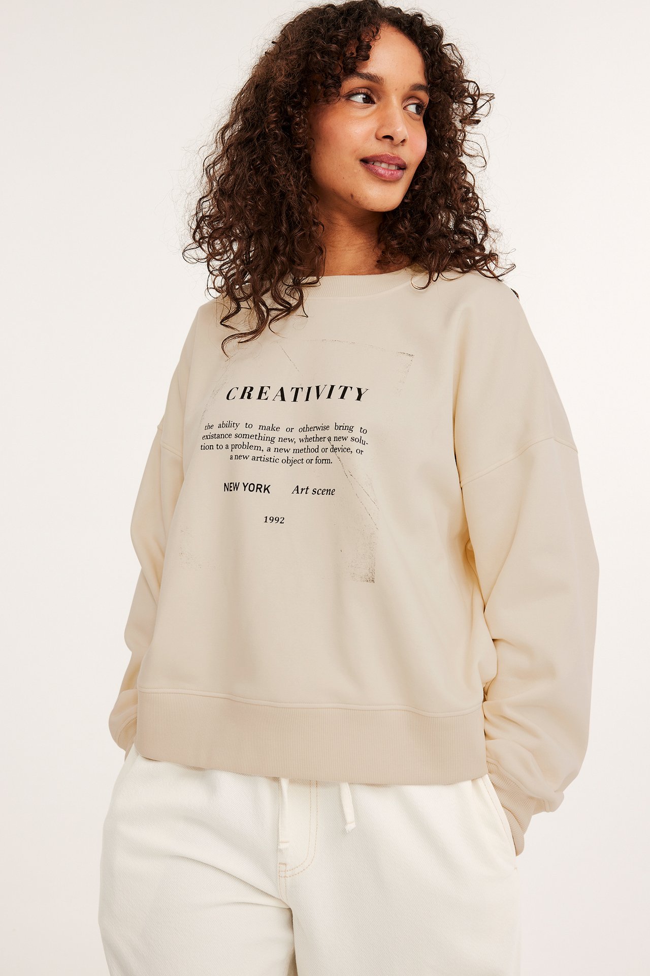 Frontbilde av en kvinne i en offwhite sweatshirt med 'CREATIVITY'-trykk og hvite joggebukser.
