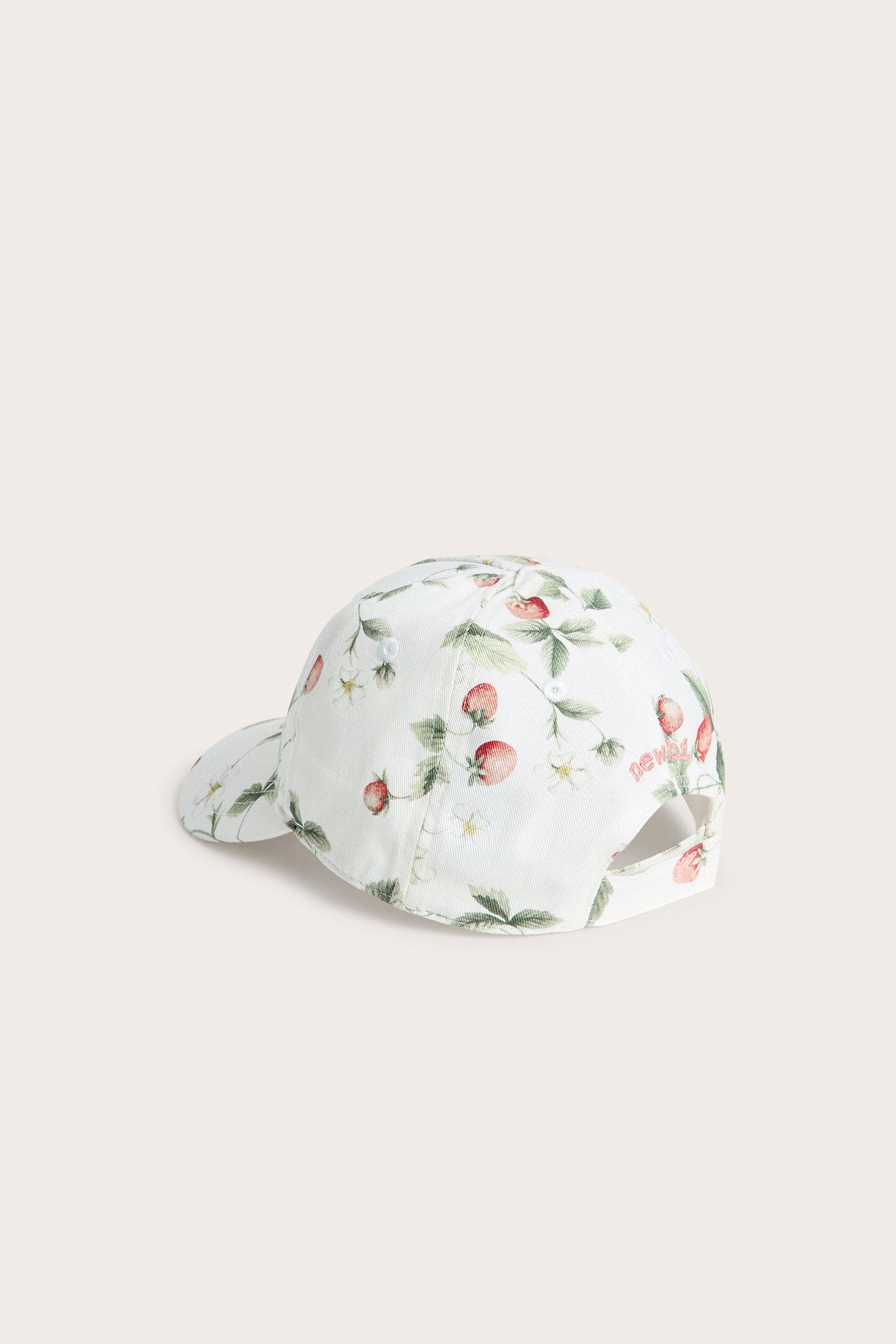 Bakside av en offwhite barnekeps med rødt jordbær- og blomstermønster, med borrelåsfeste.