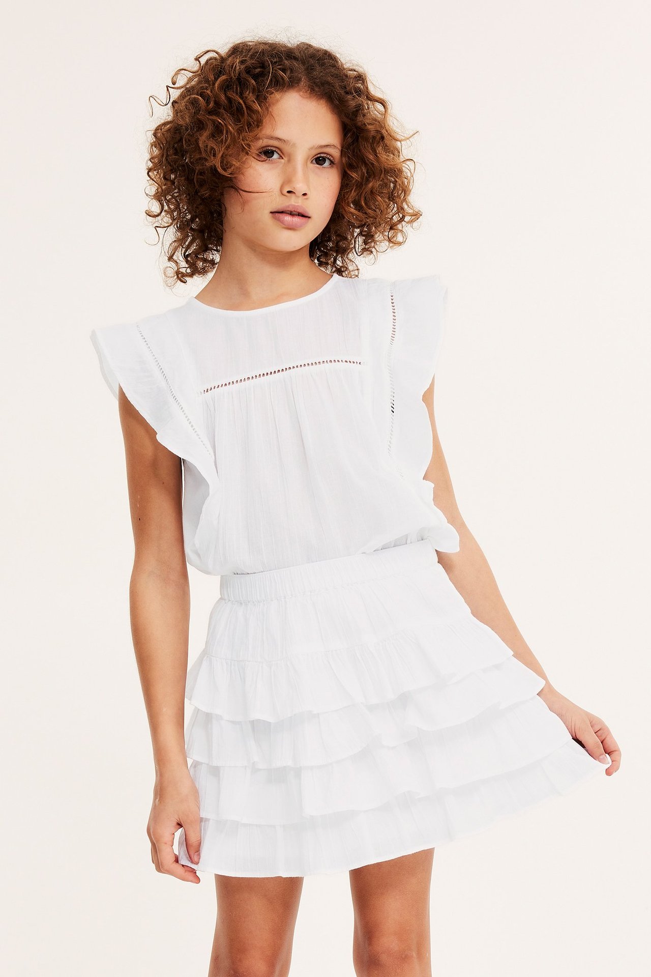 Frontvisning: Barn i hvit ermeløs bomullsbluse med volangermer, blondedetaljer og hvitt volangskjørt.