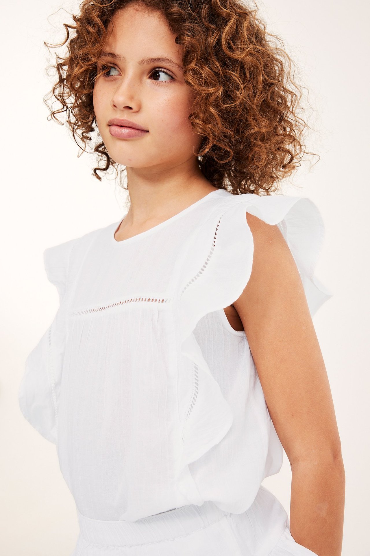 Nærbilde av et barn i hvit ermeløs bomullsbluse med volanger og blondedetaljer.