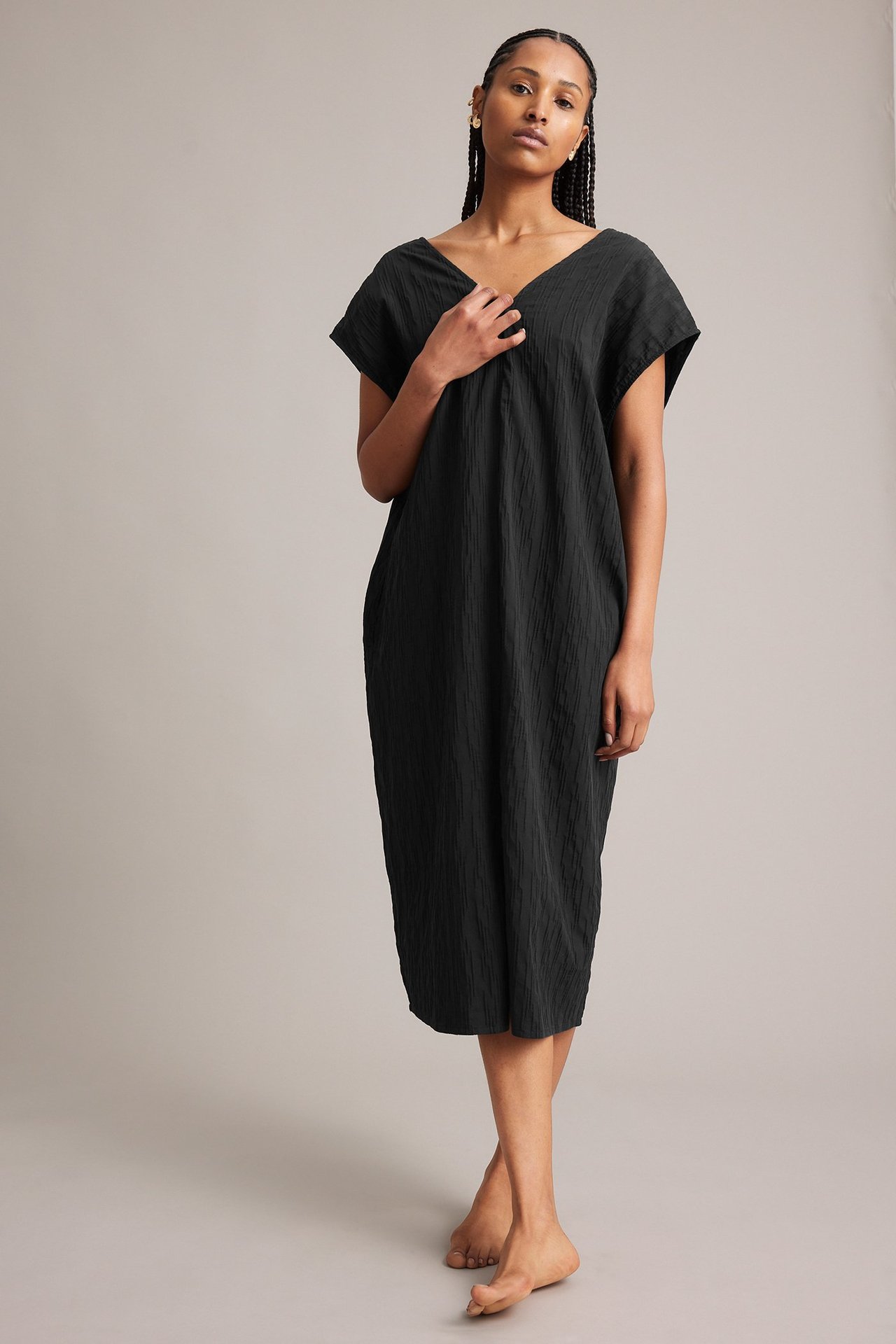 Frontutsikt: Kvinne i en svart krøllet bomullsblandingskjole med V-hals og små gulløreringer.