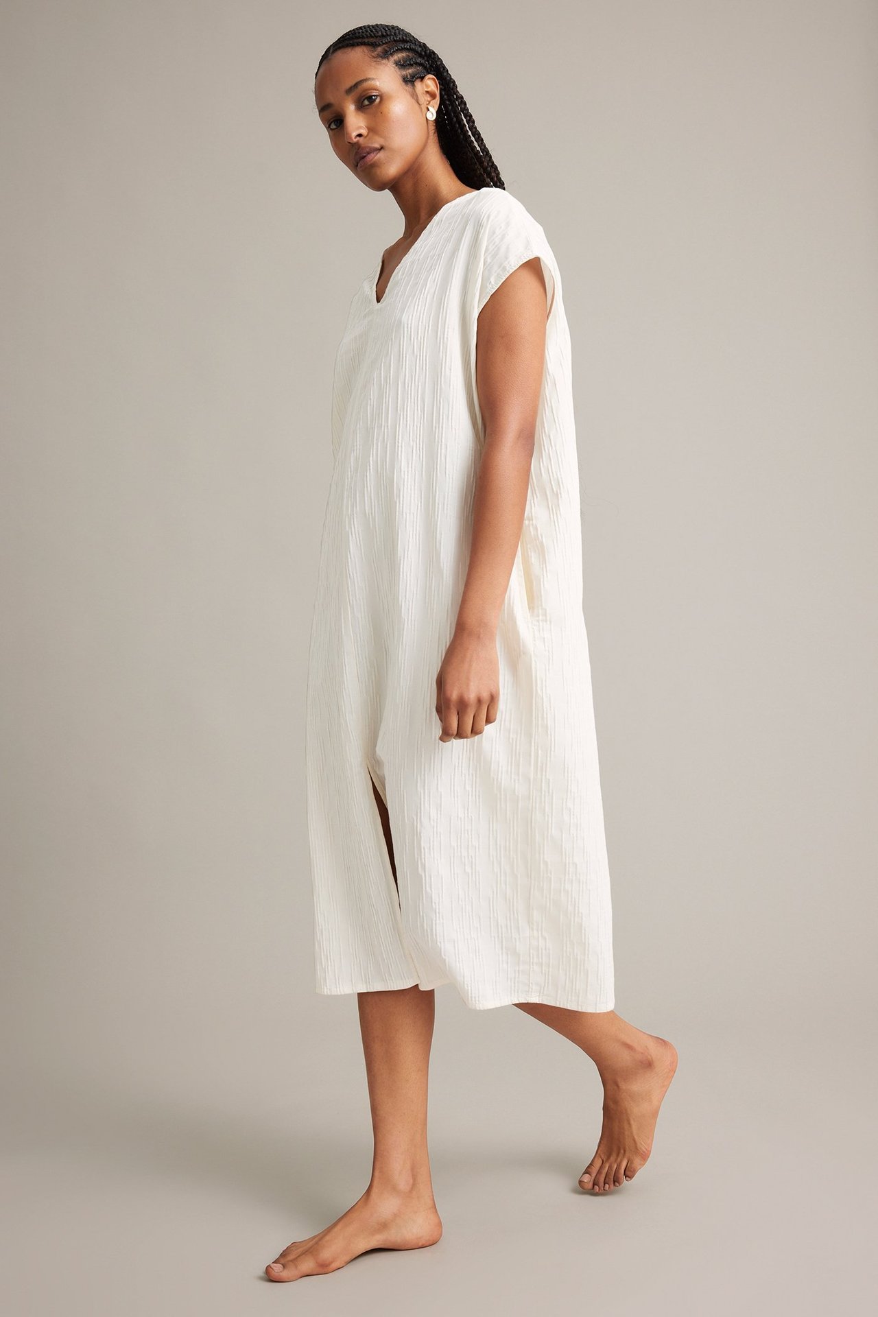 Sidevisning av kvinne i løs hvit krøllet V-hals kjole med sidesplitter og små øredobber.
