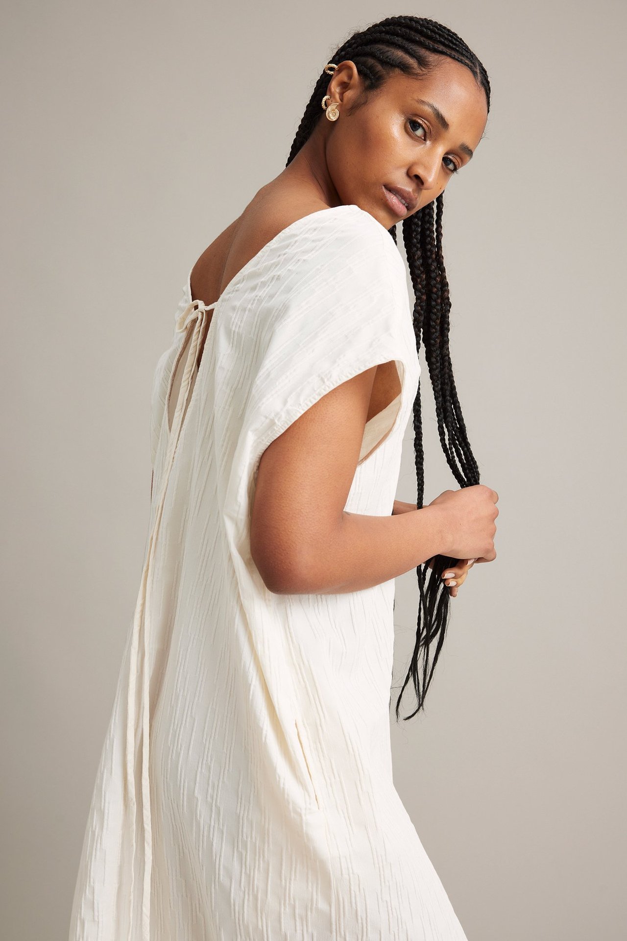 Sidebilde av kvinne i hvit krøllet bomullsblandingskjole med knytebånd bak og gullfargede øreringer.