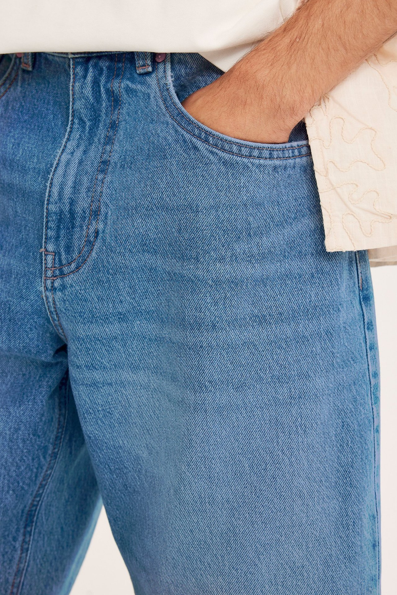 Lähikuva edestä miehestä, jolla on löysät siniset denimshortsit, käsi taskussa, vaalean kuvioidun paidan päällä.