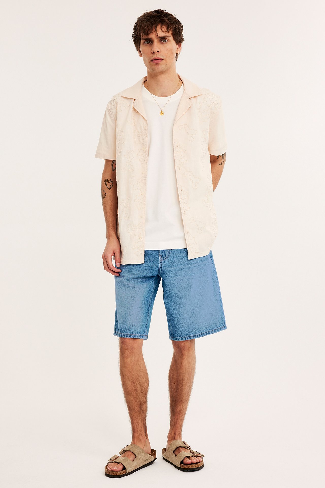 Frontvy av en man i lösa ljusblå jeansshorts, vit t-shirt, beige mönstrad skjorta och beiga sandaler.