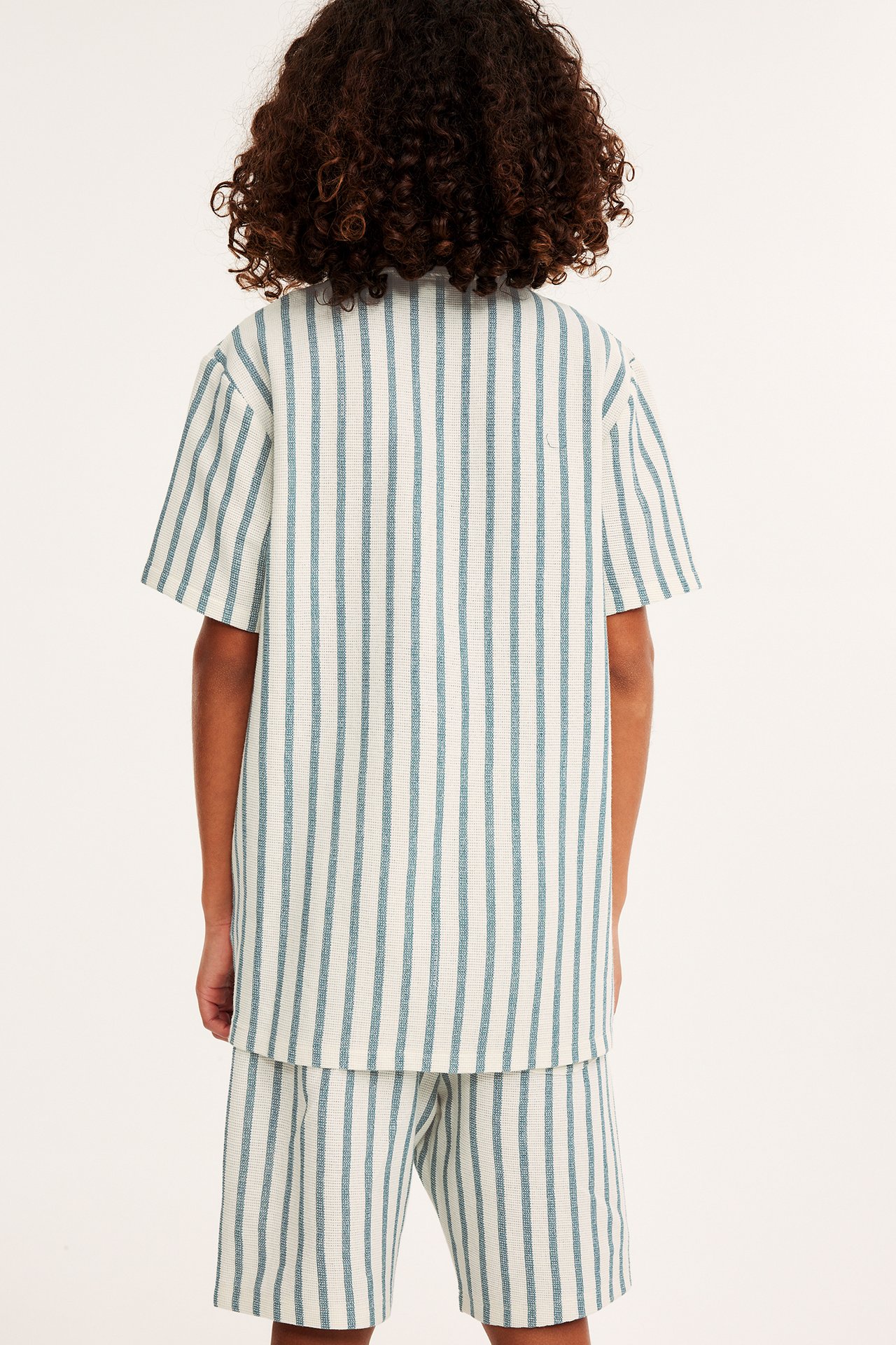Bakifrån: ett barn i en offwhite och blårandig kortärmad skjorta med matchande shorts.