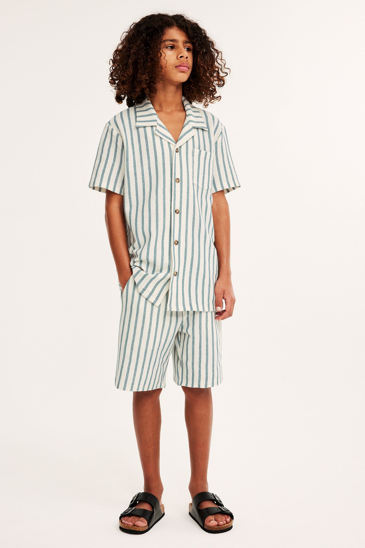 Frontvy av ett barn i offwhite och blårandig kortärmad skjorta, matchande shorts och svarta sandaler.