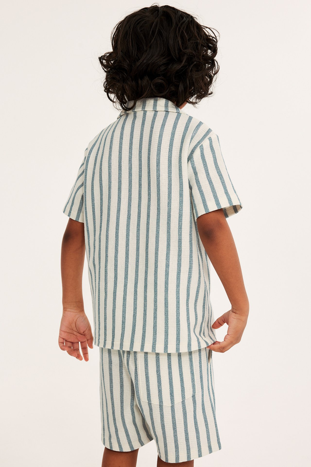 Bakifrån, ett barn i en offwhite och blårandig kortärmad jerseyskjorta med matchande shorts.