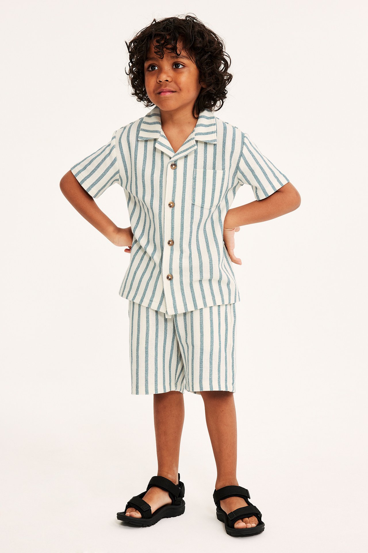 Framifrån: barn i offwhite, blårandig kortärmad skjorta, matchande shorts och svarta sandaler.