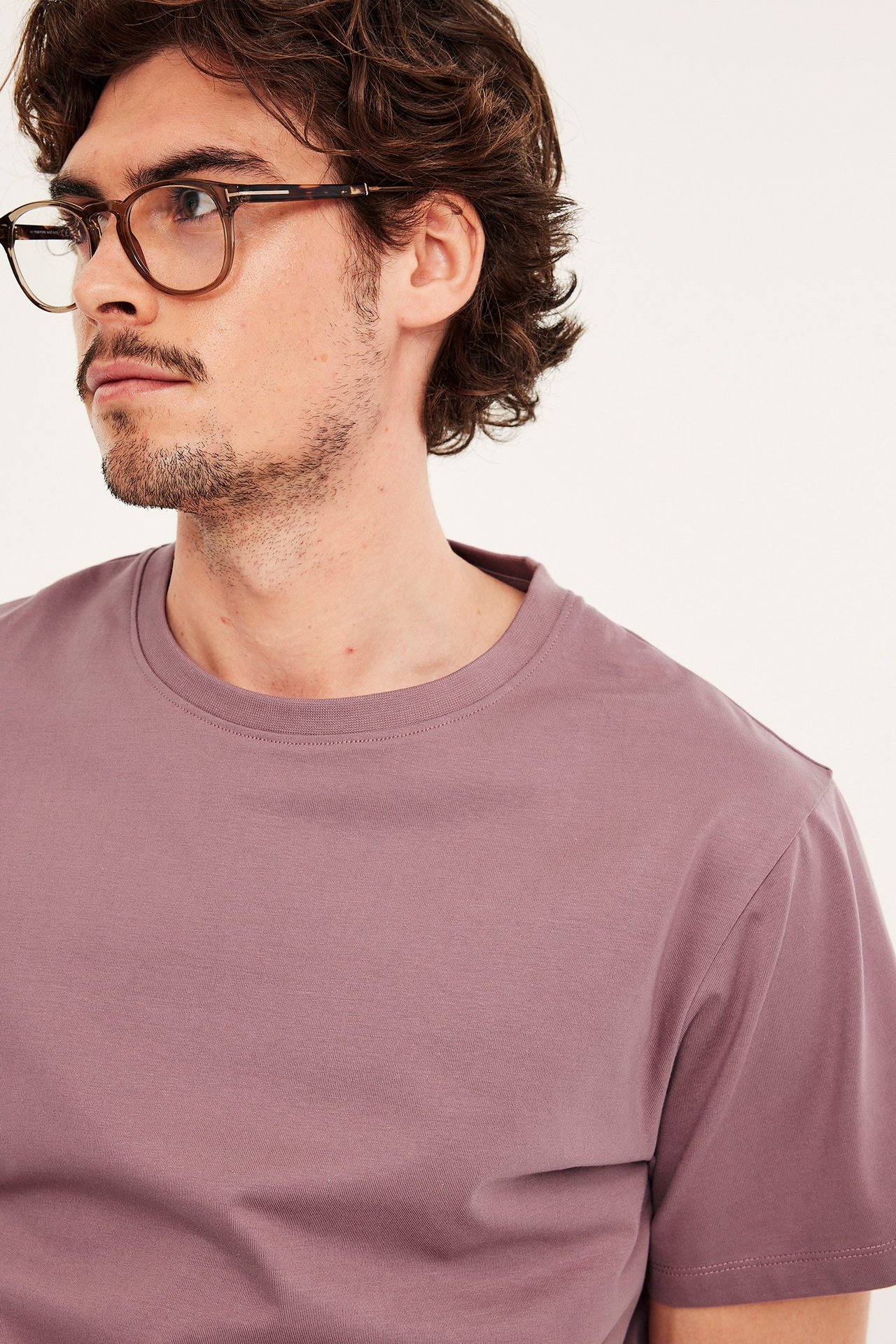 Lähikuva miehestä, jolla on pölyisen vaaleanpunainen pyöreäkauluksinen puuvillapaita ja ruskeat silmälasit.