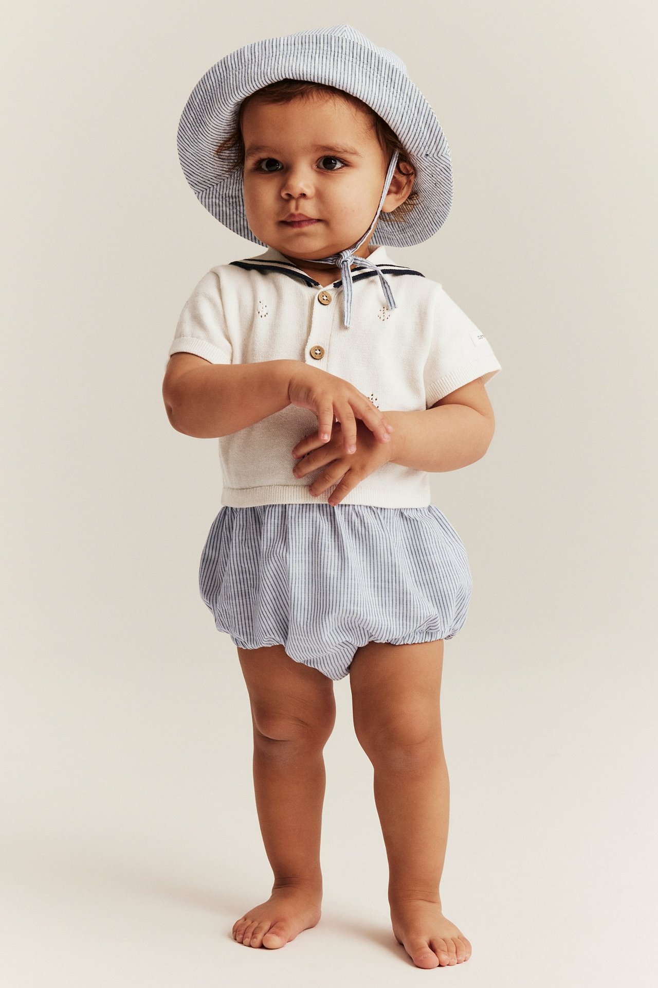 Vauva yllään luonnonvalkoinen neulehaalari merimieskauluksella, raidallinen alaosa ja raidallinen hattu.