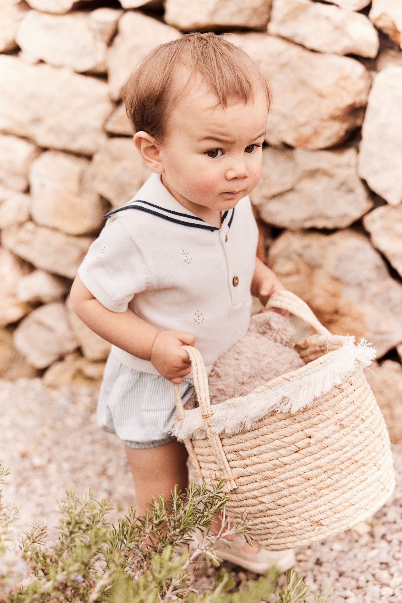Vauva yllään luonnonvalkoinen neulottu pointelle-haalari, merimieskaulus, raidallinen alaosa, pitää olkikoria.