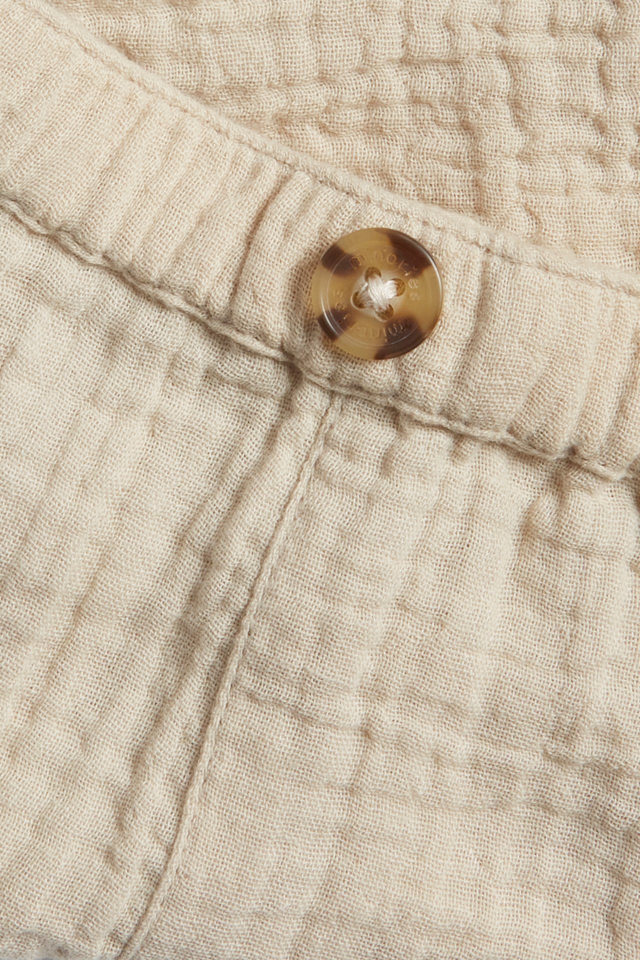 Nærbilde av lysebeige vevde musselin babyshorts med elastisk linning og en brun pynteknapp.