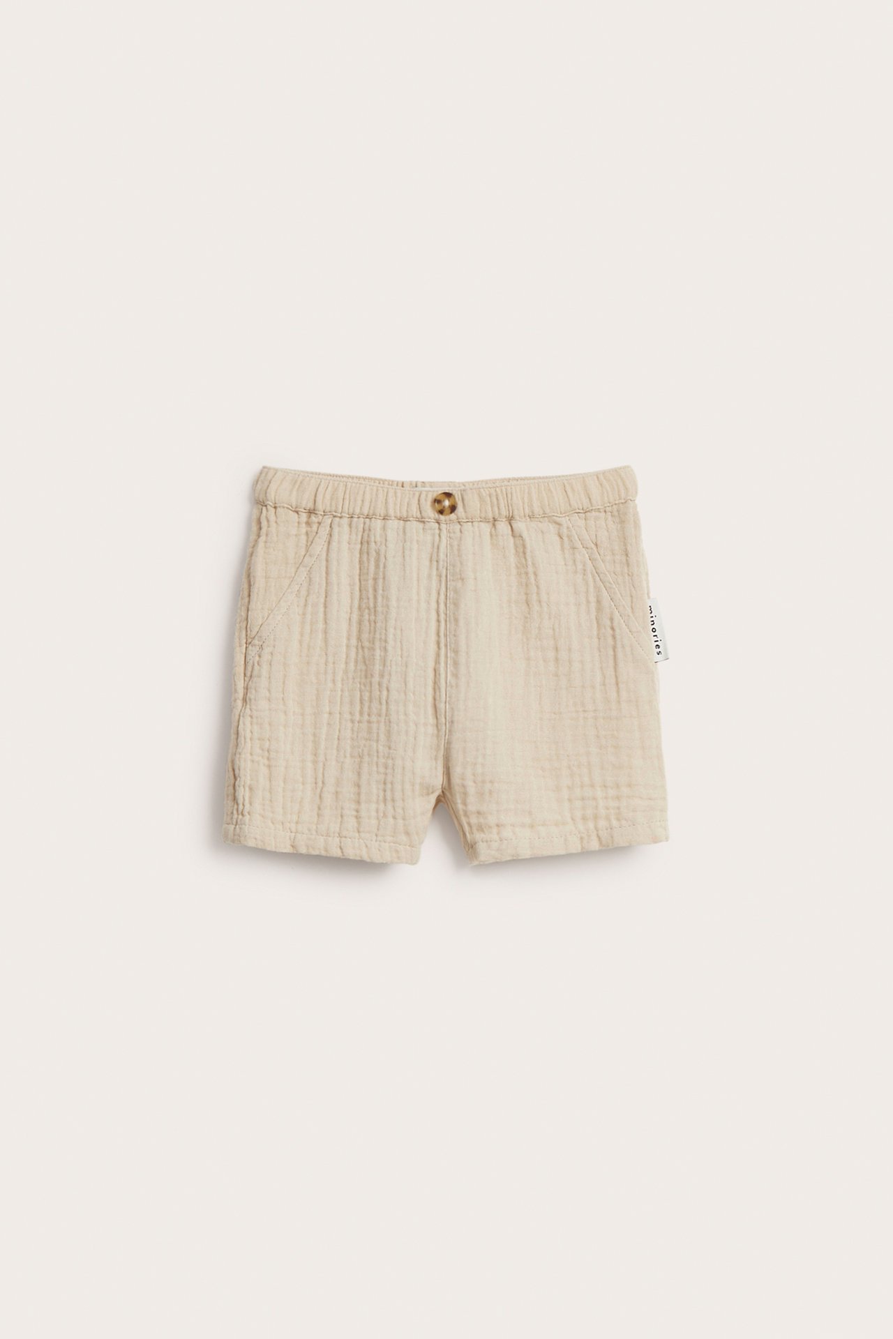 Frontvisning av lysebeige babyshorts i vevd musselin med elastisk midje og en pynteknapp.