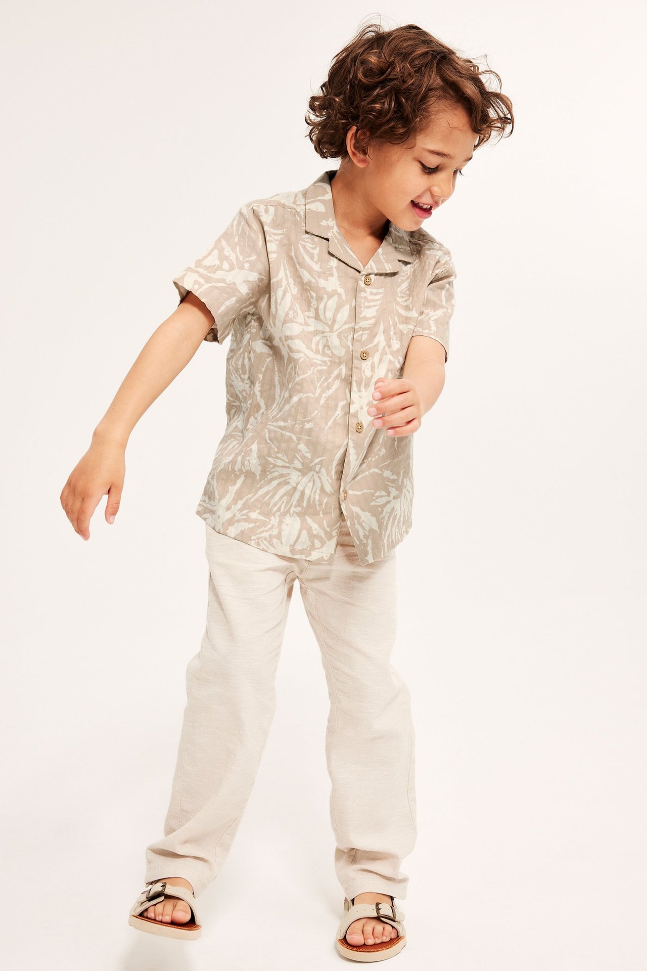 Frontvy av ett barn i en ljusbeige mönstrad bäckebölja resortskjorta, ljusbeige byxor och sandaler.