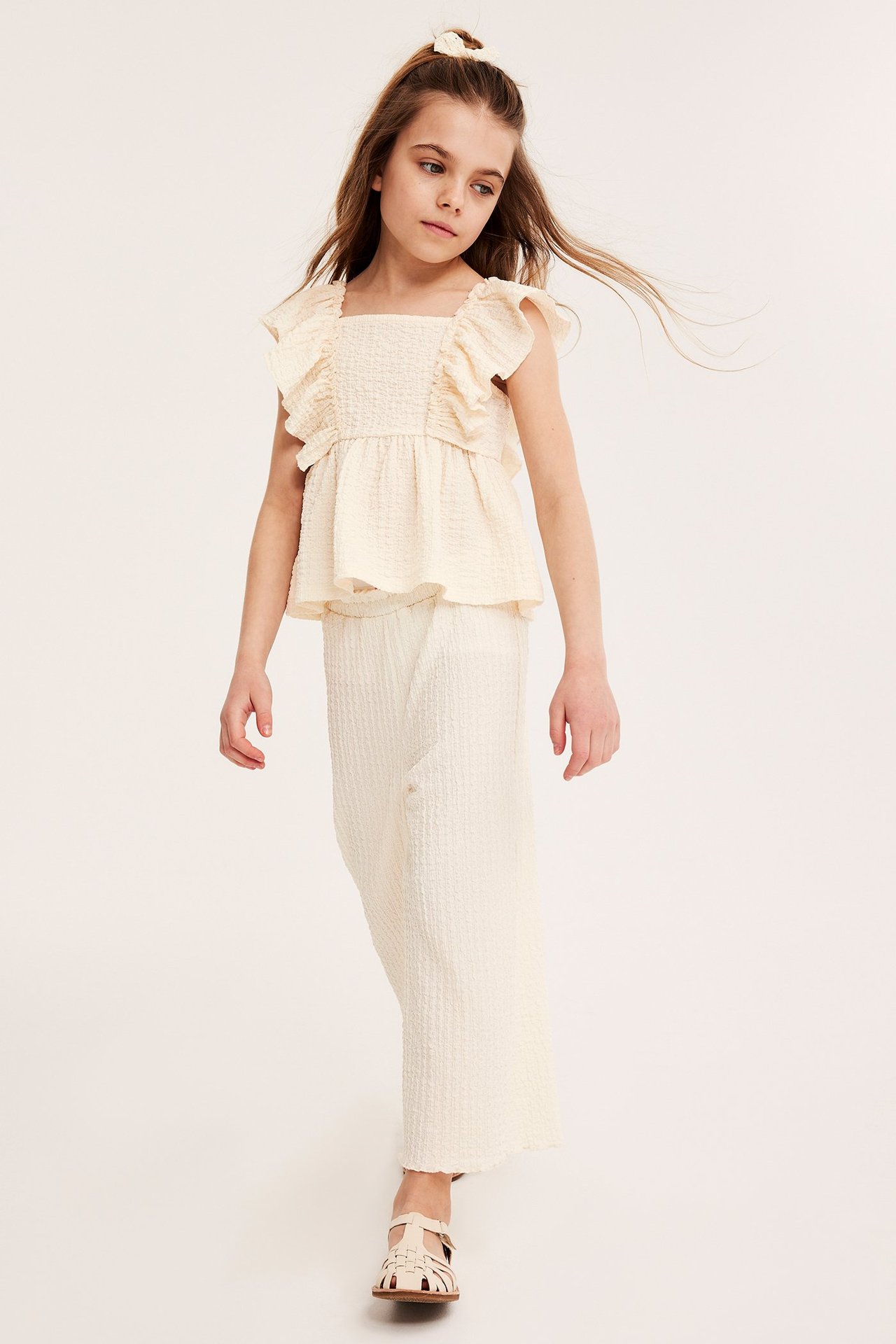 Widok z przodu dziecka w kremowym, strukturalnym topie z falbanami, pasujących szerokich spodniach i jasnobeżowych sandałach.