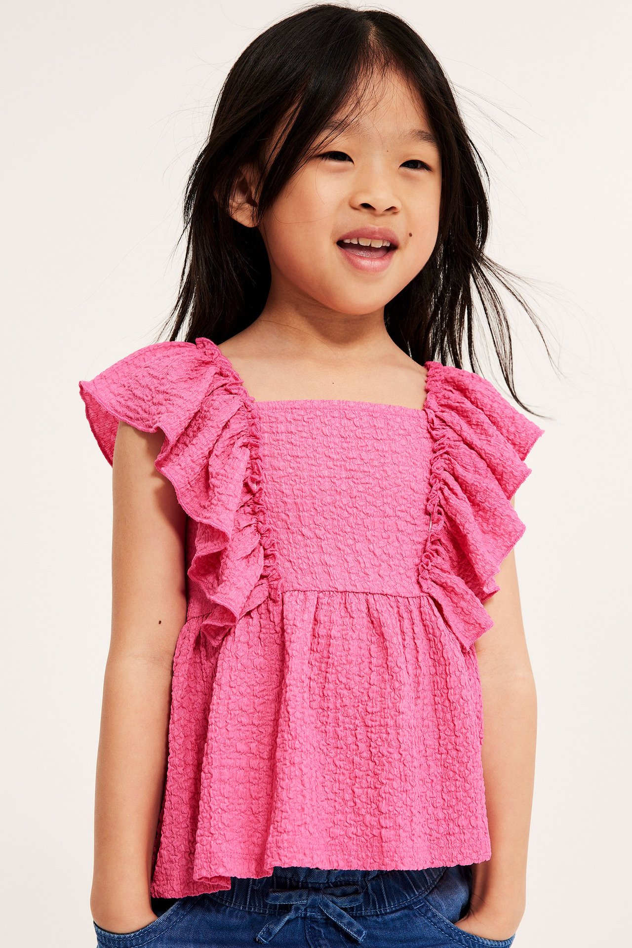 Widok z przodu: Dziecko w różowym topie z teksturowanej dzianiny z dużymi falbanami i niebieskich spodniach.
